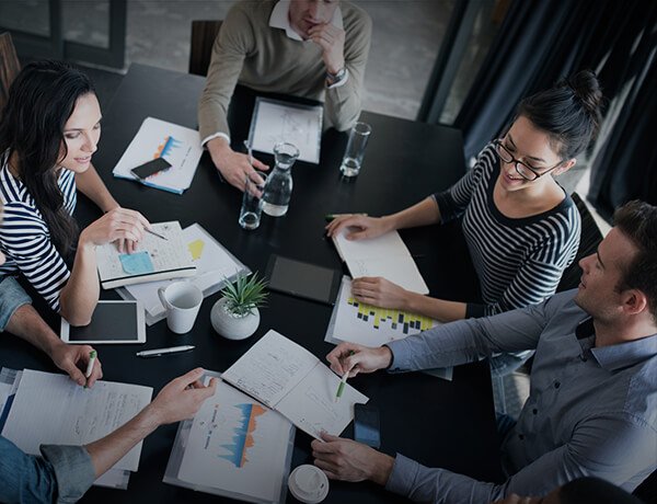 A group of five people in a business meeting sitting around a table with documents and charts, engaging in discussion and taking notes.