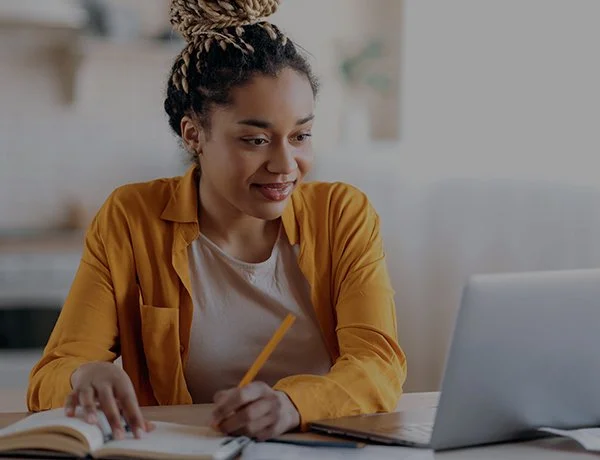A woman with curly hair in a bun, wearing a yellow shirt over a white top, sitting at a desk with a pen, notebook, and a laptop in a bright room.