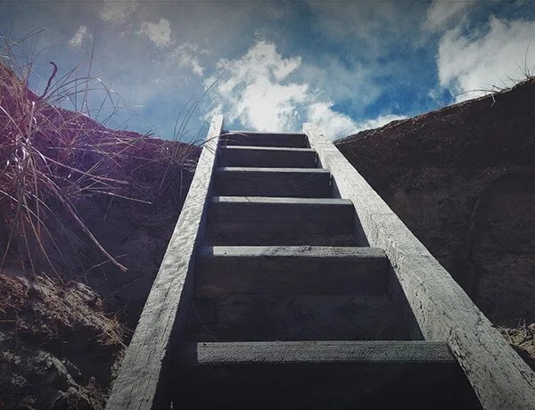 View looking up a wooden ladder outdoors with blue sky and clouds in the background, surrounded by dirt and grass.