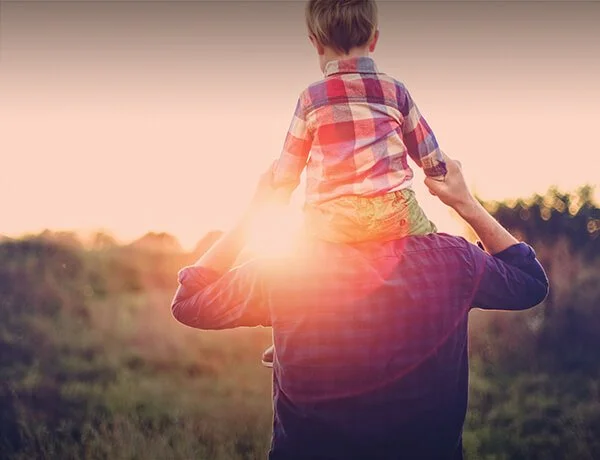 Child sitting on adult's shoulders during sunset in a grassy field.