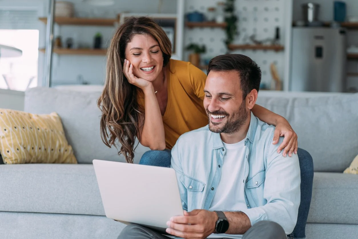 A man sitting on the floor using a laptop with a woman leaning over his shoulder, smiling, in a bright living room.