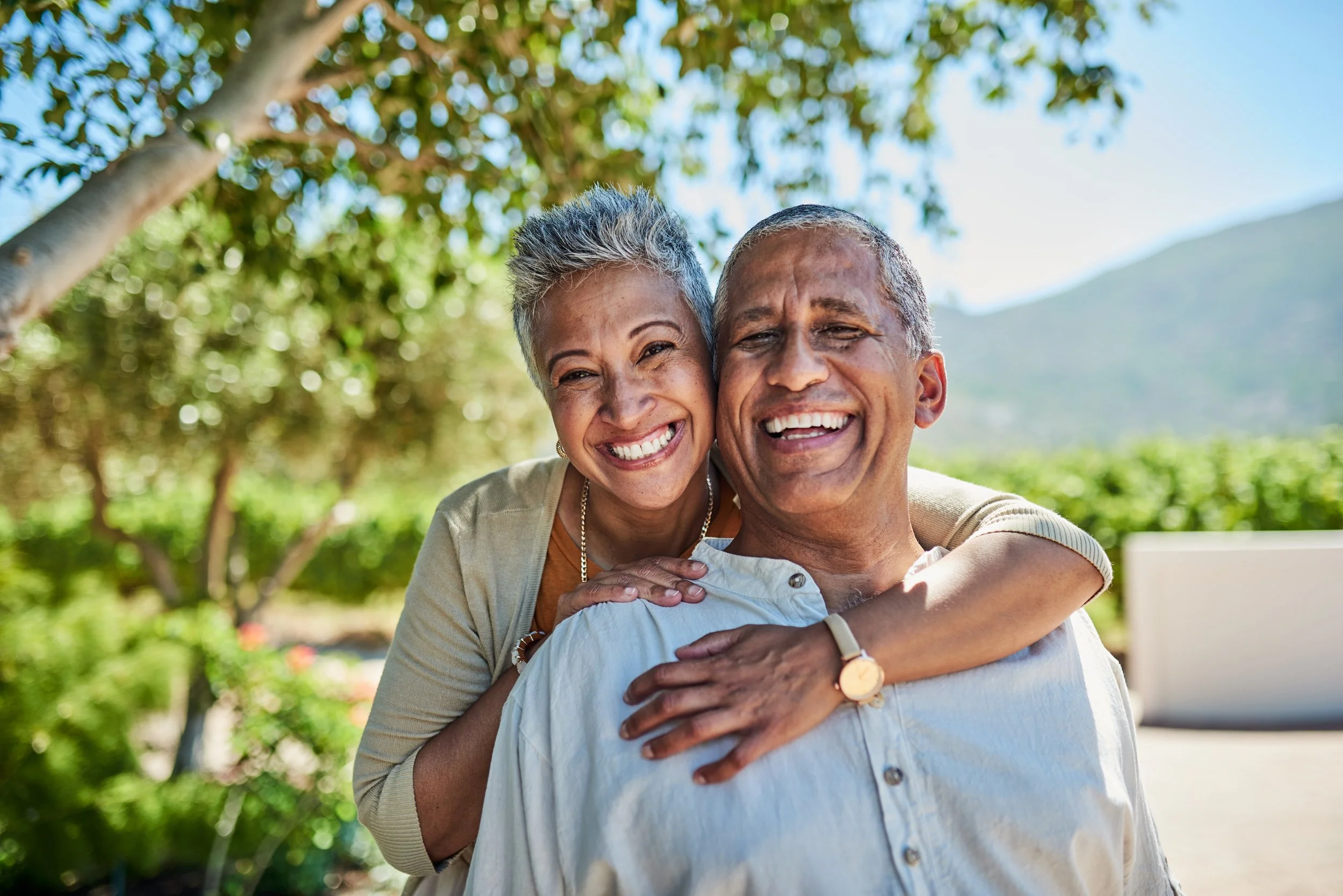 A happy older couple smiling and embracing outdoors with trees and mountains in the background.