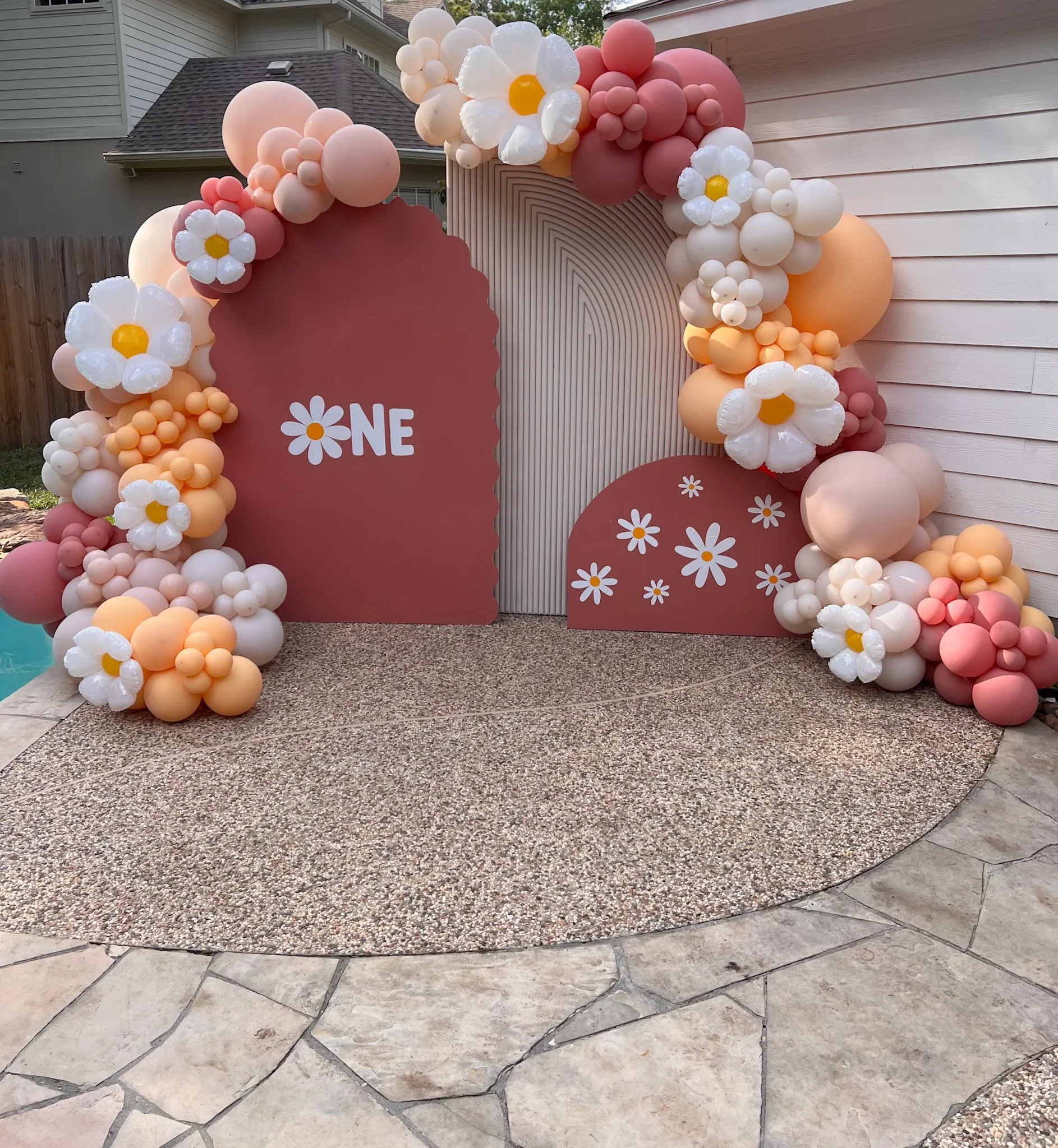 Decorative floral balloon arch with pink, peach, white, and coral balloons, featuring large flower-shaped balloons with white petals and yellow centers, surrounding a pale pink backdrop with the word "ONE" and daisies, set up outdoors on a paved surf