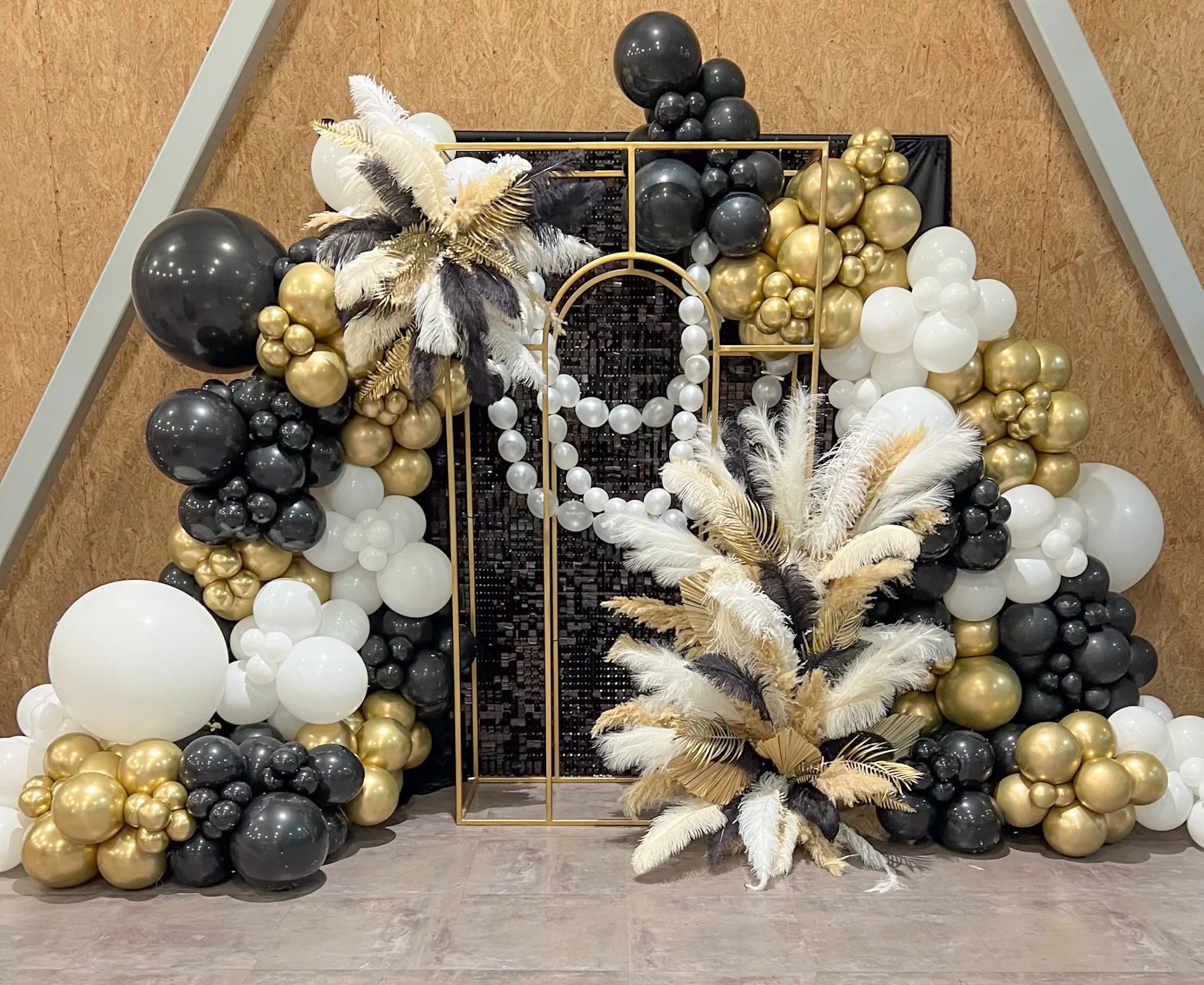 Decorative balloon arch with black, white, and gold balloons, adorned with white and black feathers and gold leaves, set against a black sequin backdrop.
