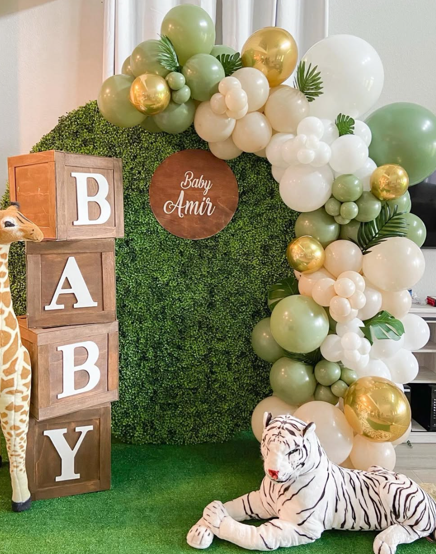 Decorative backdrop for a baby celebration with a green leafy wall, white and gold balloons, a wooden circle sign with Baby Amir written on it, and a stack of blocks spelling 'BABY'. There is a plush zebra toy in front and a plush giraffe partially v