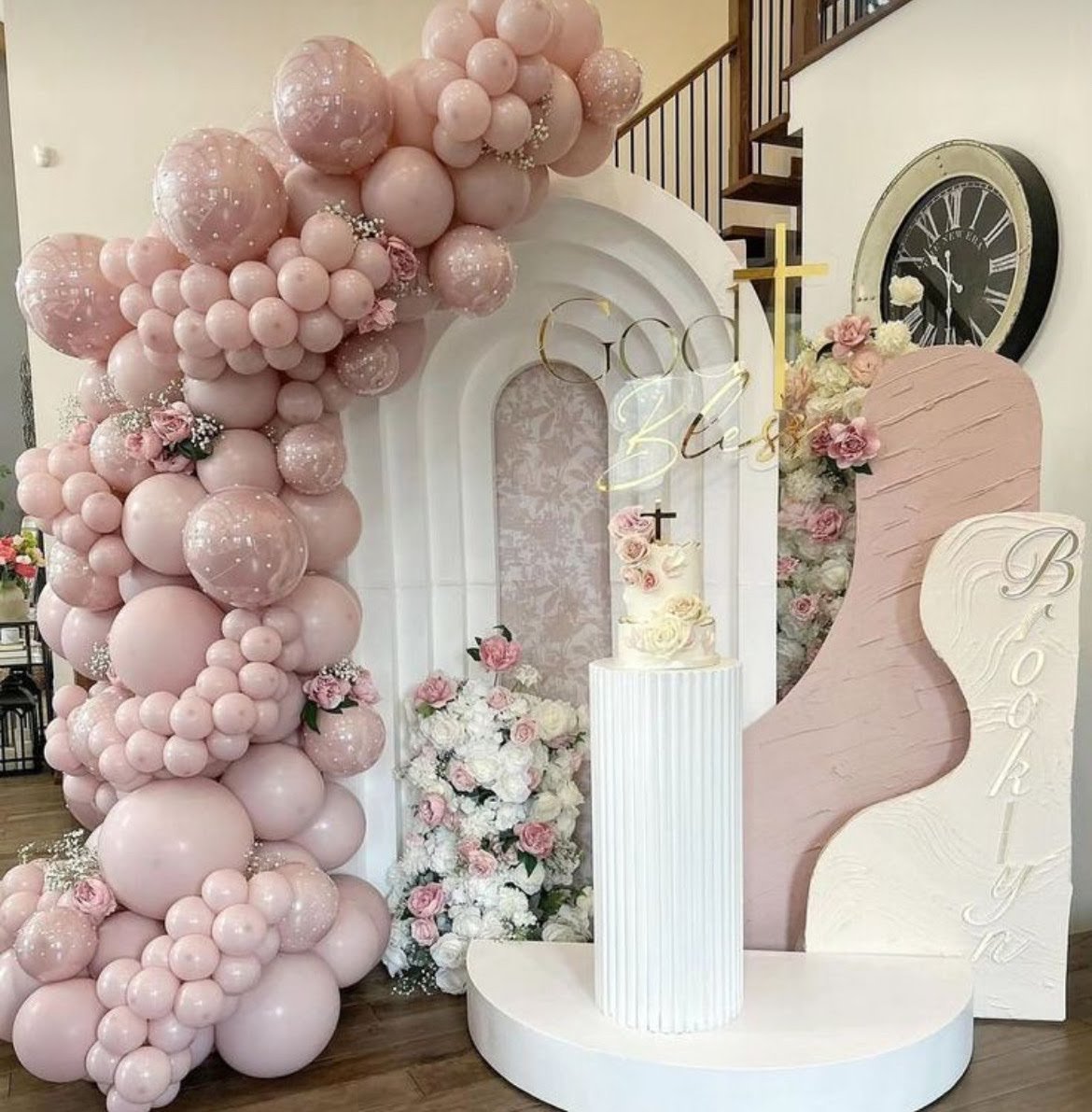Decorative event setup with pink and white balloons, flowers, a cake on a pedestal, and signs that say 'God Bless' and 'Baby Girl' in a church or event space.