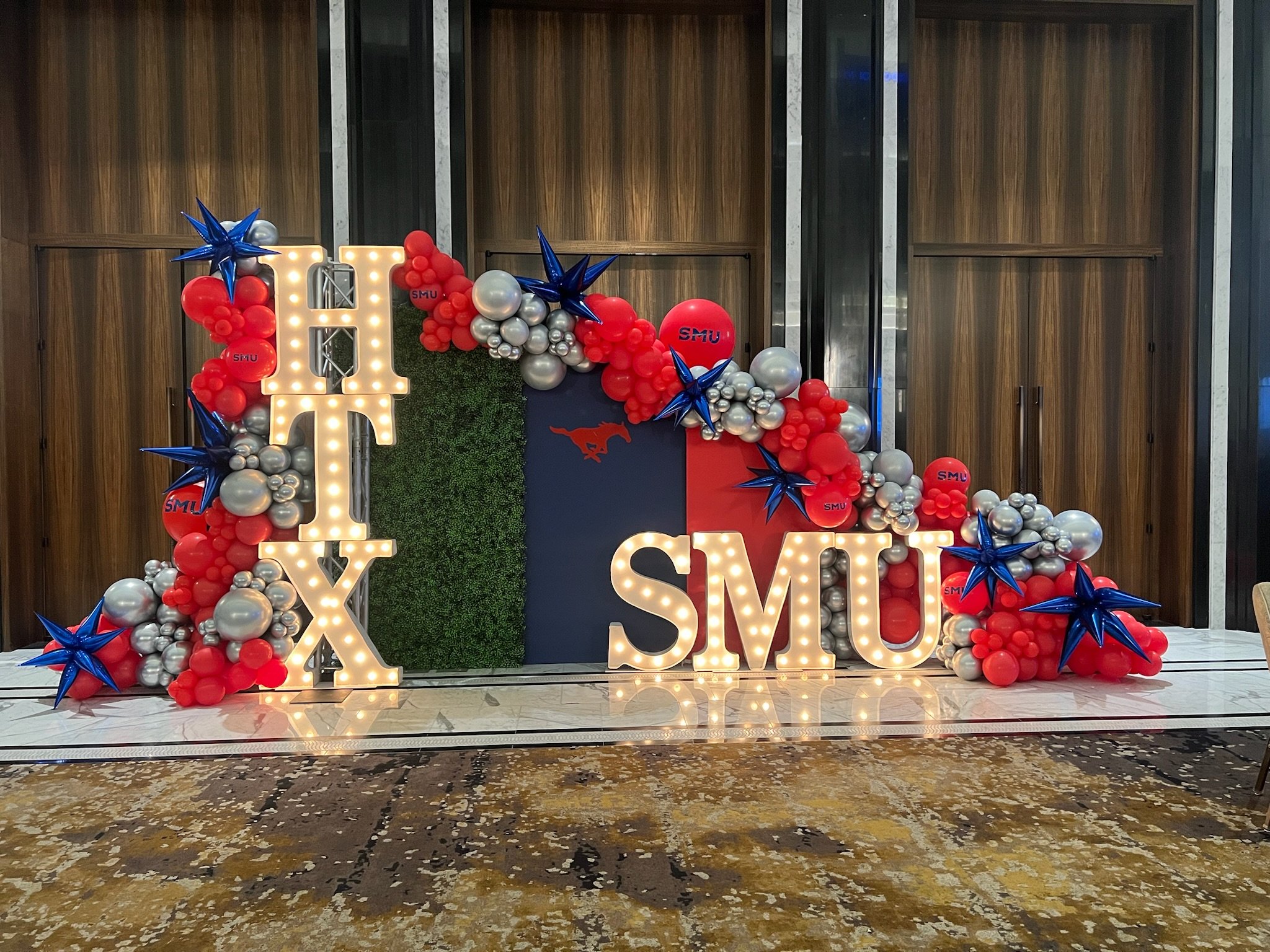 Decorative setup with illuminated letters spelling 'FOX' and 'SMU', surrounded by red, silver, and blue balloons with star and Mustang designs, on a marble floor in front of wooden wall panels.