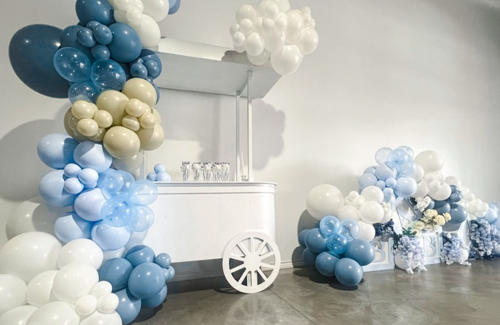 Decorated backdrop with blue, white, and cream-colored balloons arranged around a white cart with a canopy, along with floral arrangements.