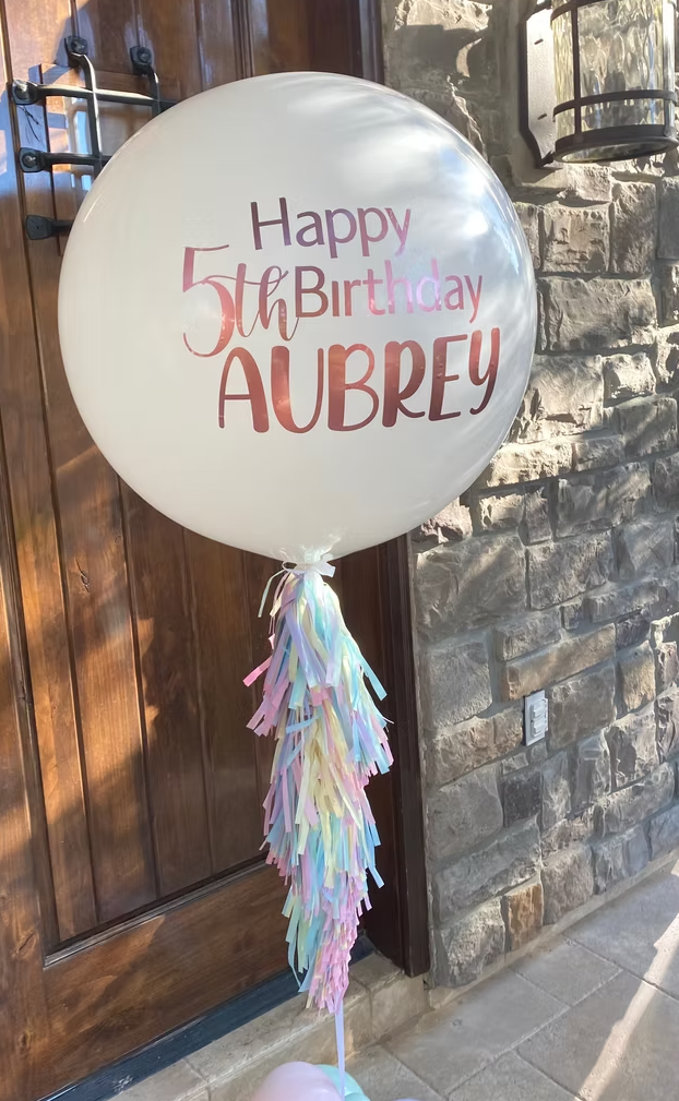 Balloon with the words "Happy 5th Birthday AUBREY" printed on it, decorated with pastel-colored tassels hanging from the bottom, placed near a stone wall and wooden door.