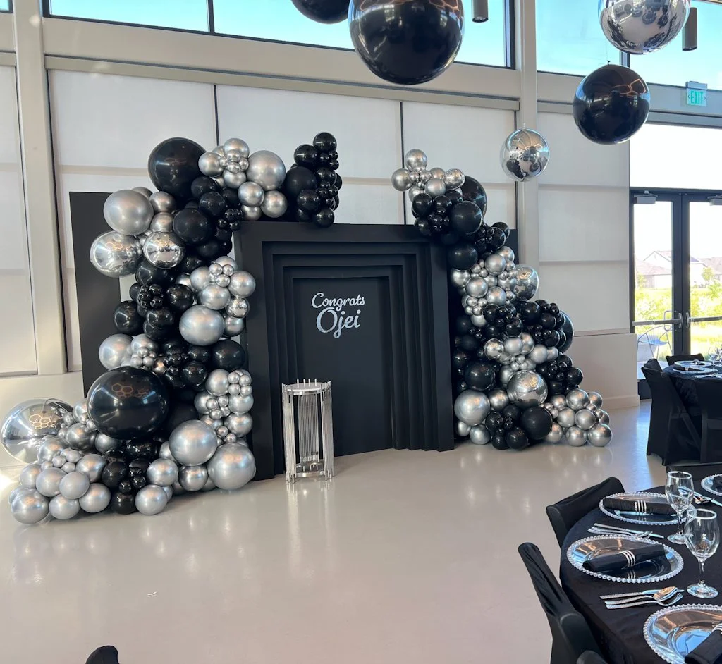 Elegant balloon decoration in black, silver, and chrome surrounds a black photo backdrop with the words "Congrats Ojei" written on it, in a modern event space with hanging black and silver balloons.