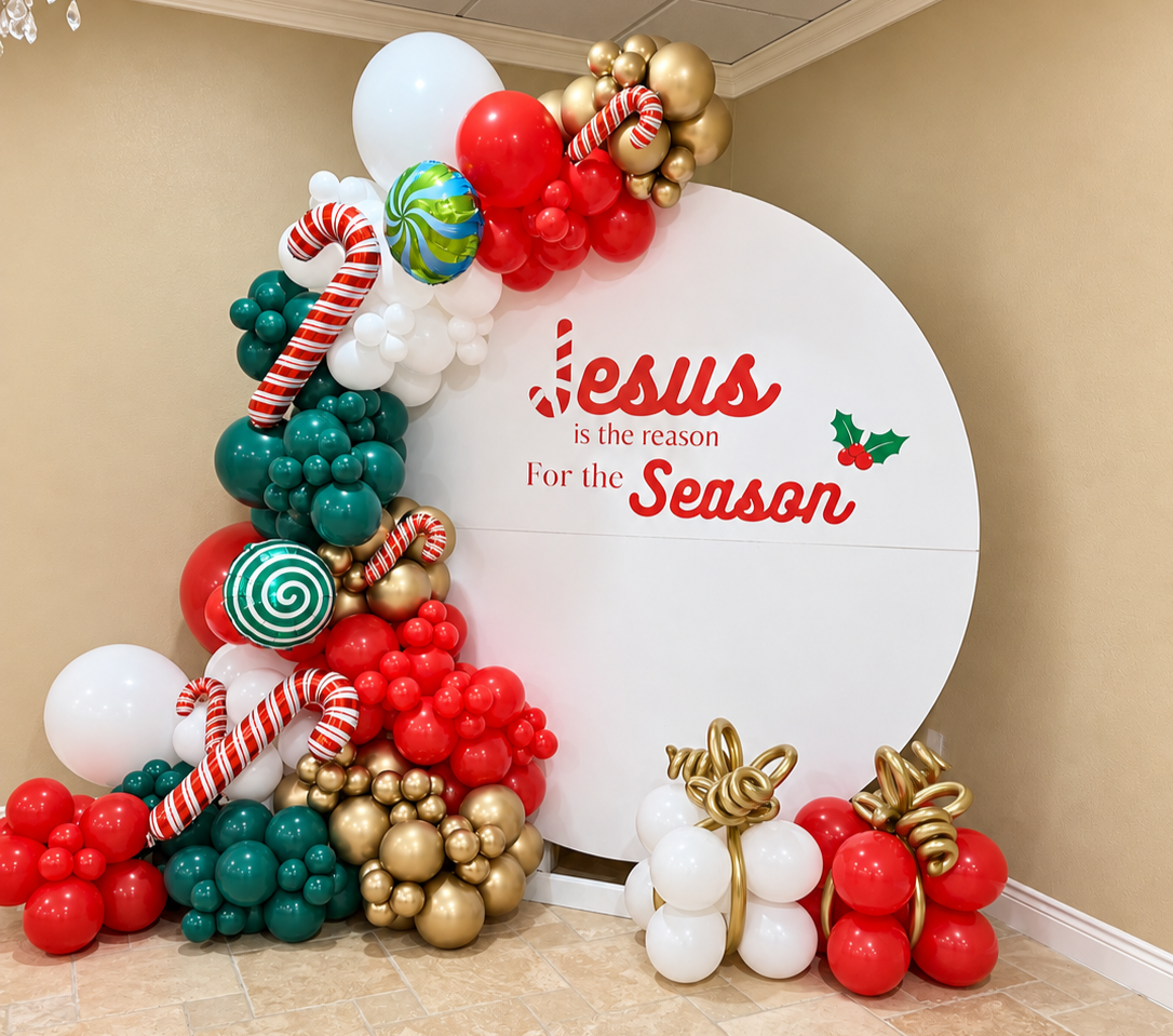 White round backdrop with a “Jesus is the reason for the season” message, red, green and gold balloons, candy canes, and balloon gifts.