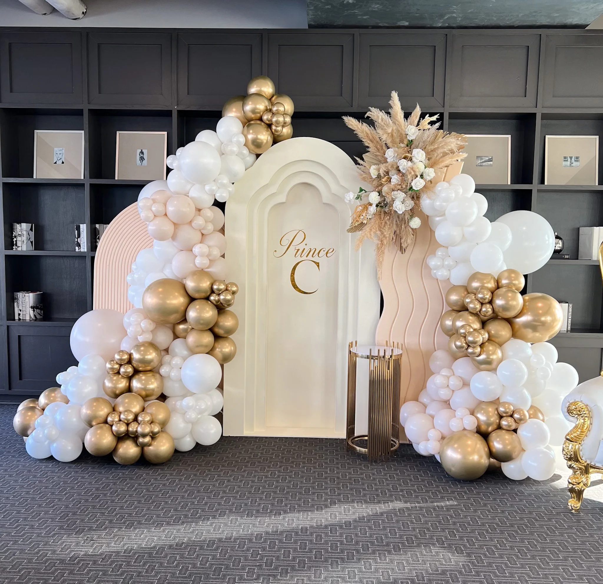 Decorative backdrop with balloon arrangements in white, gold, and blush pink, featuring a floral bouquet with dried pampas grass and white roses, and a sign with the text 'Prince C' in gold. The backdrop has a wavy design with a large arch in the center.