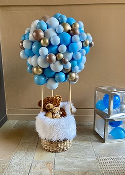 Decorative display with a basket filled with stuffed animals and a balloon arrangement above it, featuring blue, gold, and white balloons shaped like a cloud. A small transparent box with blue balloons is nearby.