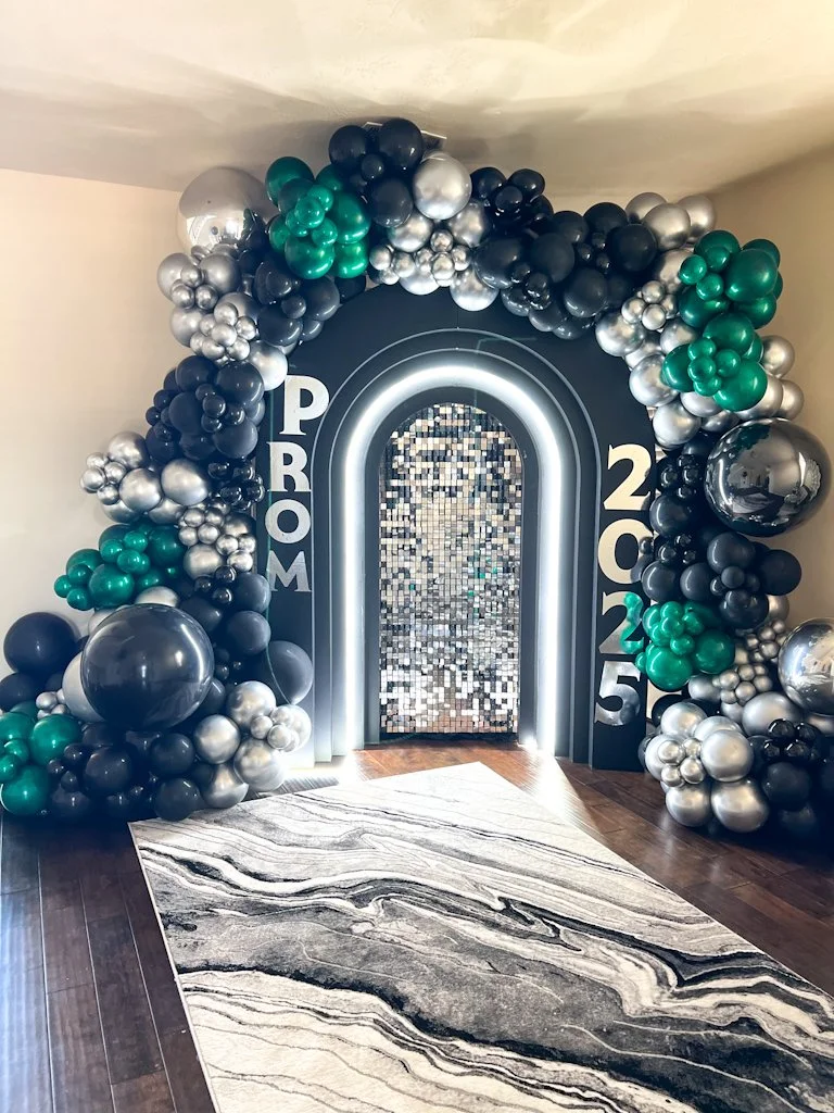 Decorative balloon arch with black, silver, and green balloons surrounding an arched doorway with a mirror mosaic and illuminated outline. The numbers '2025' and the word 'PROM' are displayed on either side of the arch, indicating a prom event in 202