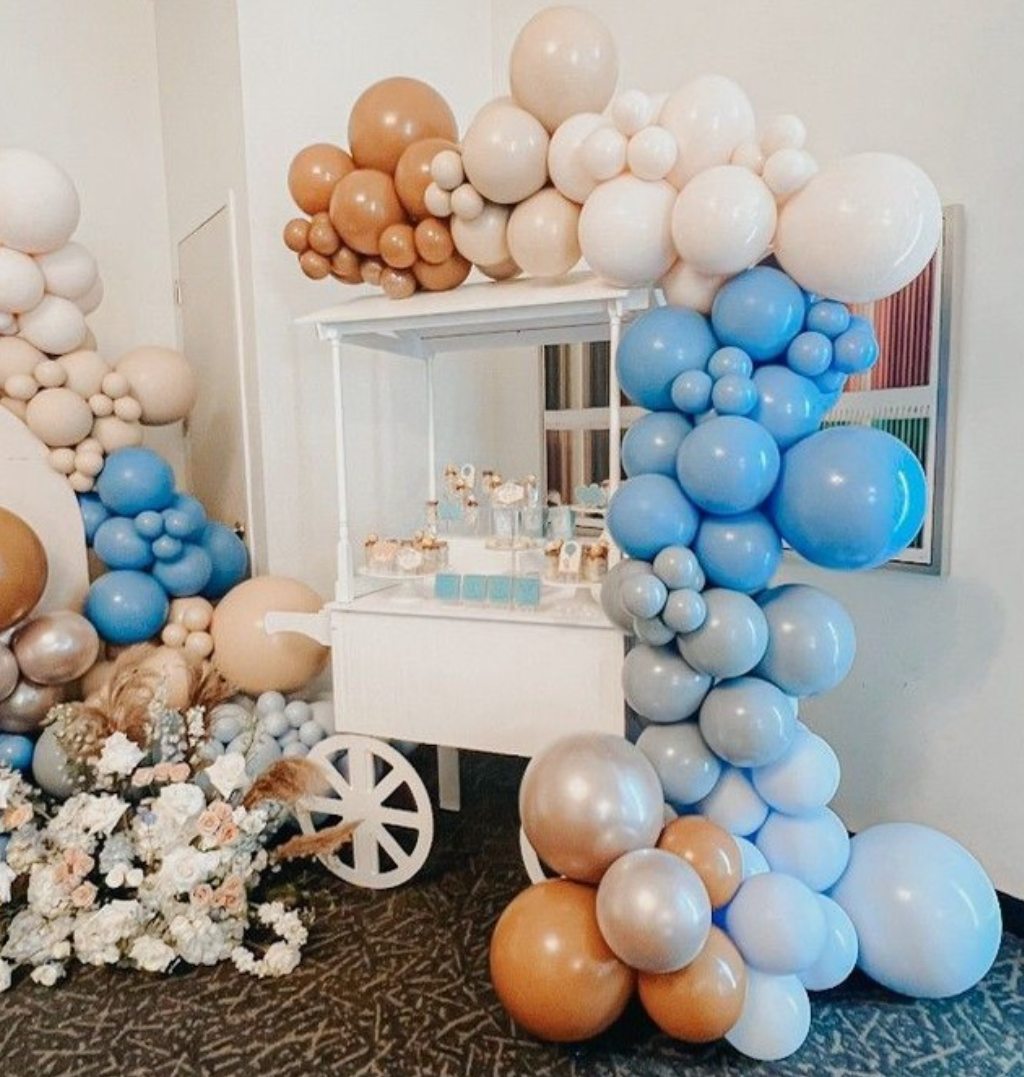 Decorative balloon arrangement with white, blue, beige, and gold balloons surrounding a white cart with desserts and surrounded by flowers.