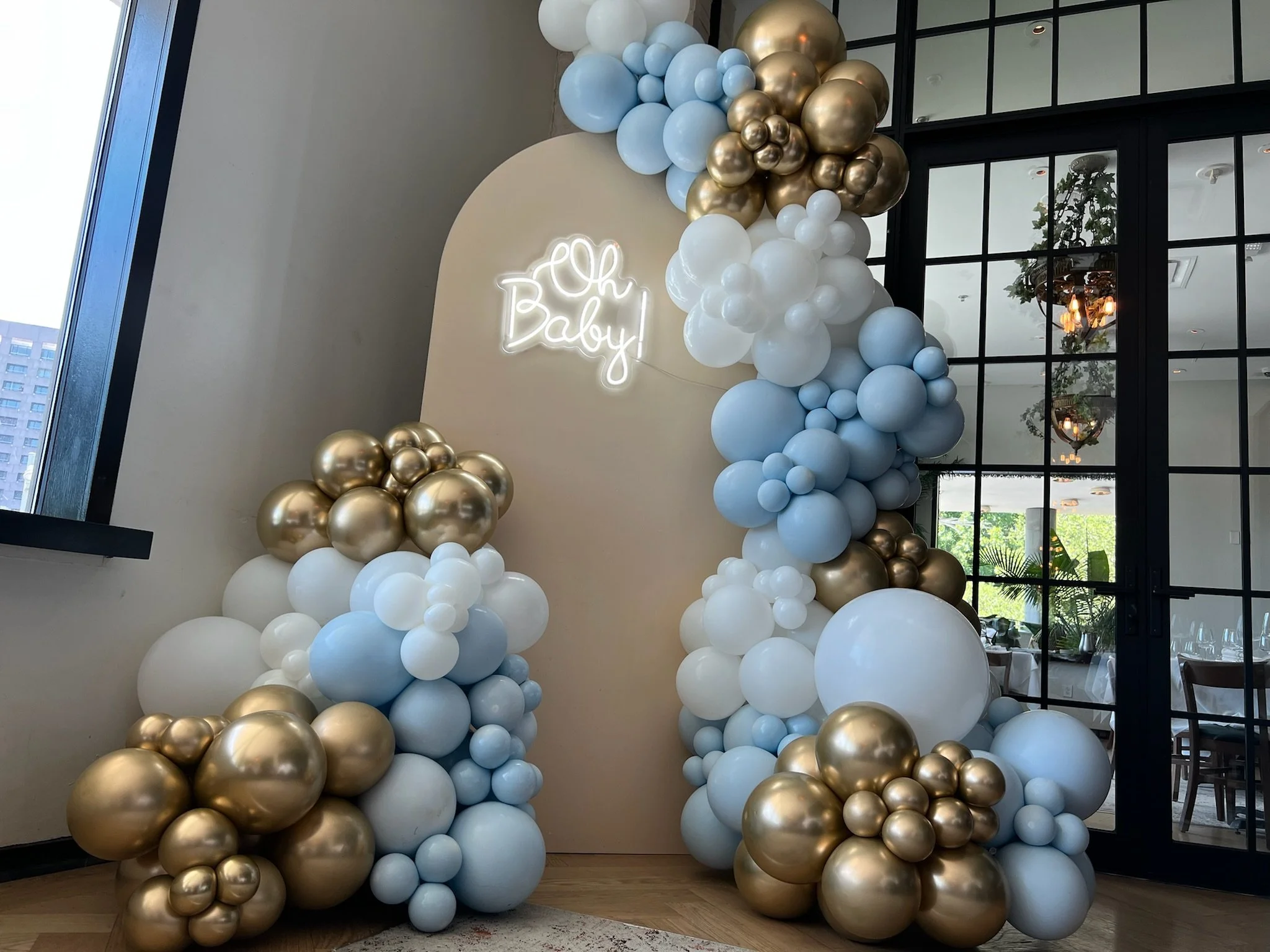 Decorative balloon arrangement with white, gold, and light blue balloons, with a neon sign saying 'Oh Baby!' in the background.