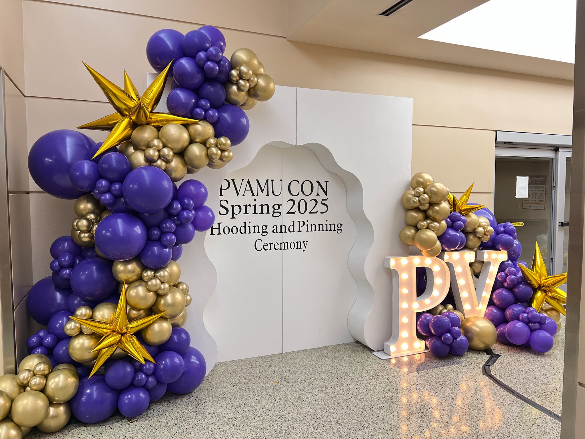 Decorative balloon arch with purple and gold balloons and gold star balloons, with a large lit-up 'PV' sign, at a graduation ceremony event.