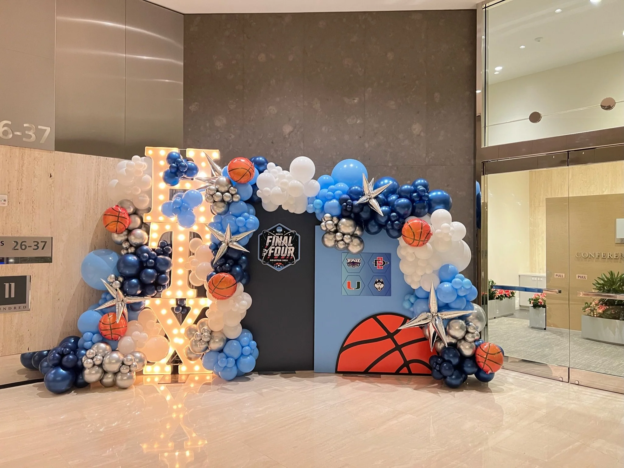 Decorative display featuring balloons in blue, white, silver, and orange, arranged around a large letter 'L' illuminated with lights, with basketball-themed and Final Four elements, in a lobby or reception area.