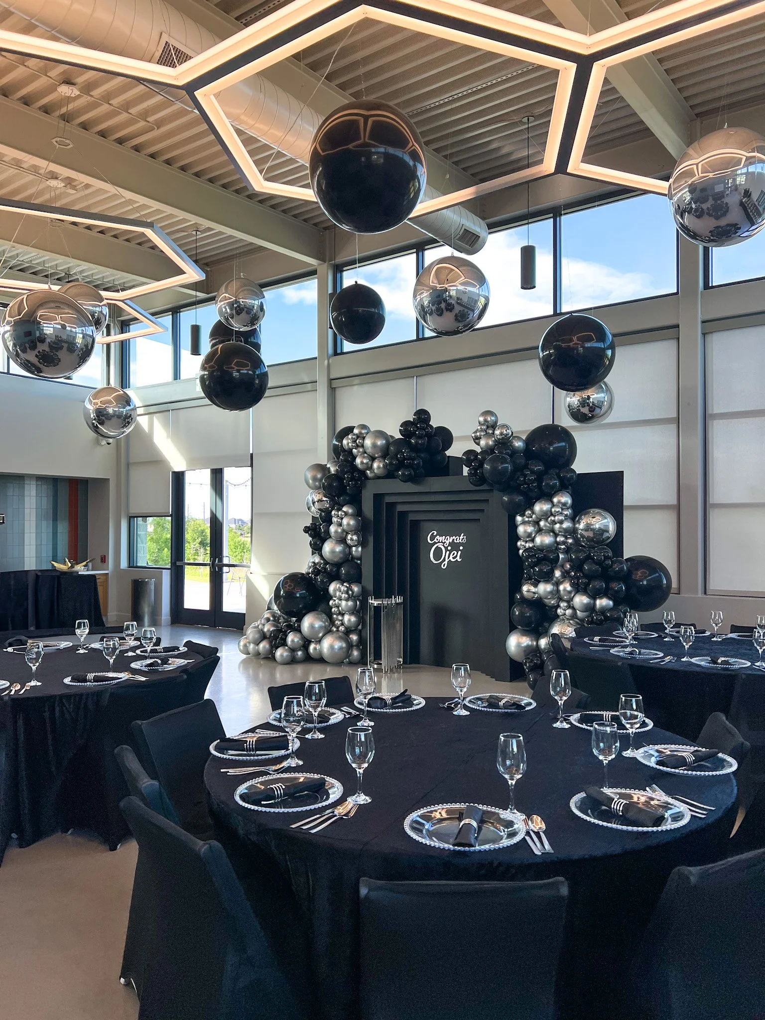 Modern decorated event space with black tablecloth-covered round tables set with glassware and napkins, black and silver balloon arrangements, black geometric backdrop with the message 'Congrats Oje,' and large black and silver hanging spherical deco