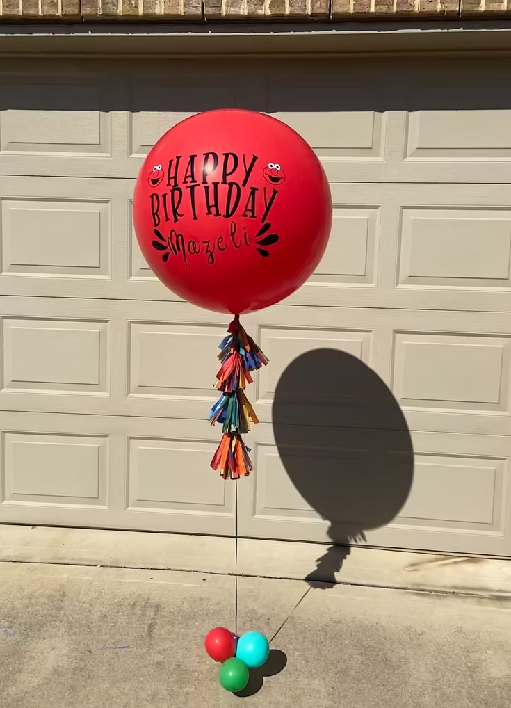 A red birthday balloon with colorful paper tassels hanging from the string, with the words "Happy Birthday Mazeli" and smiley faces printed on it, casting a shadow on a garage door.