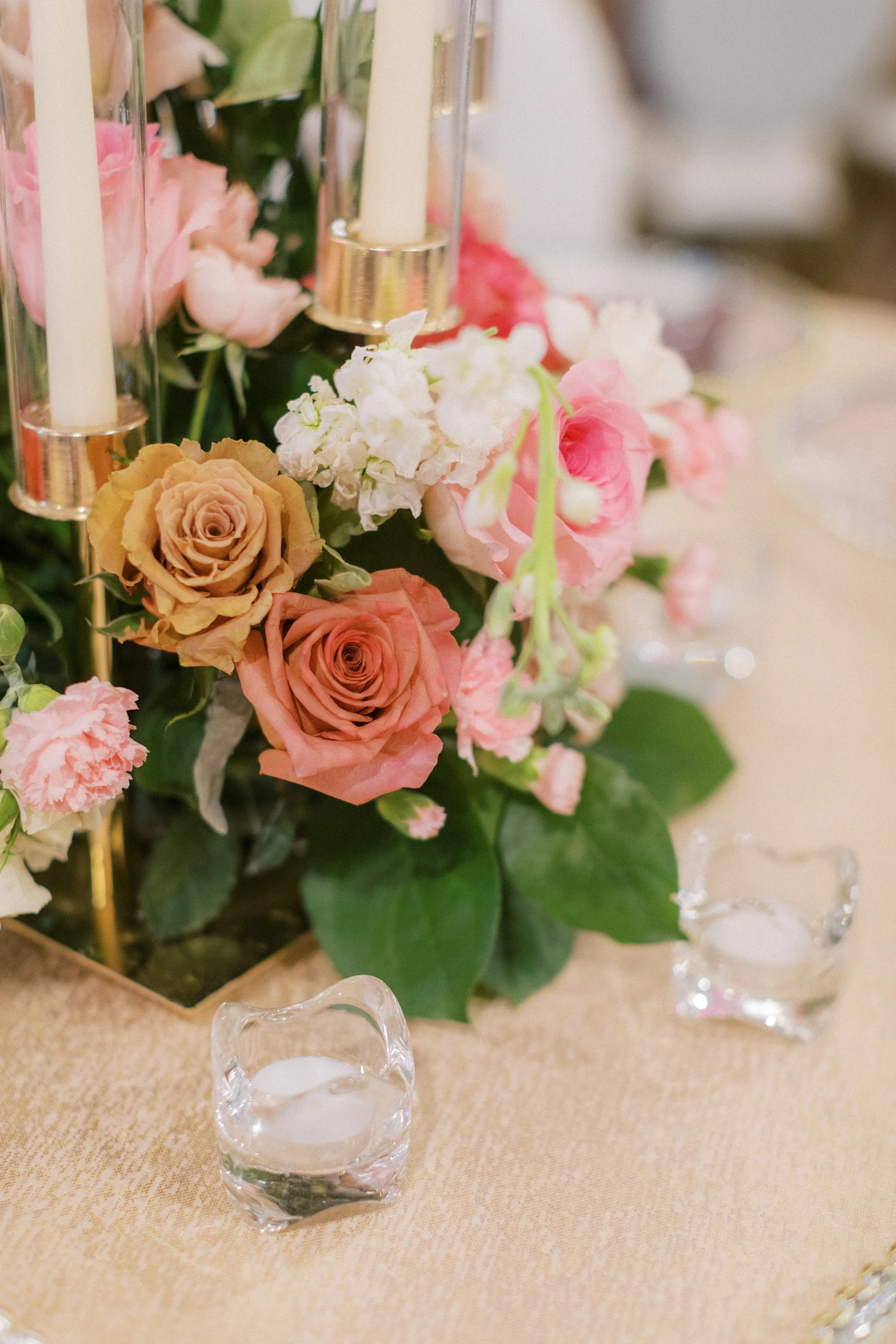 Elegant floral table centerpiece with pink, peach, and white roses, surrounded by green leaves, with glass candle holders with white candles and glass bowls filled with water on a gold tablecloth, at a formal event or wedding.