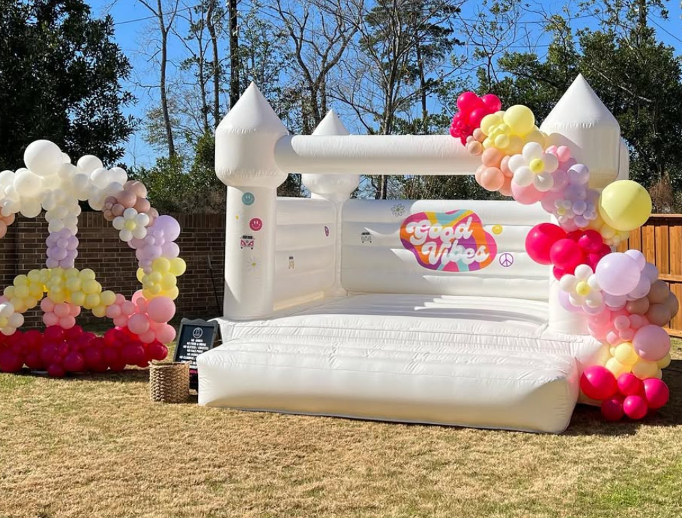 Inflatable bounce house with a pizza theme labeled 'Good Vibes' in a backyard, decorated with colorful balloon clusters in pink, purple, yellow, and white, and set against a backdrop of trees and a blue sky.