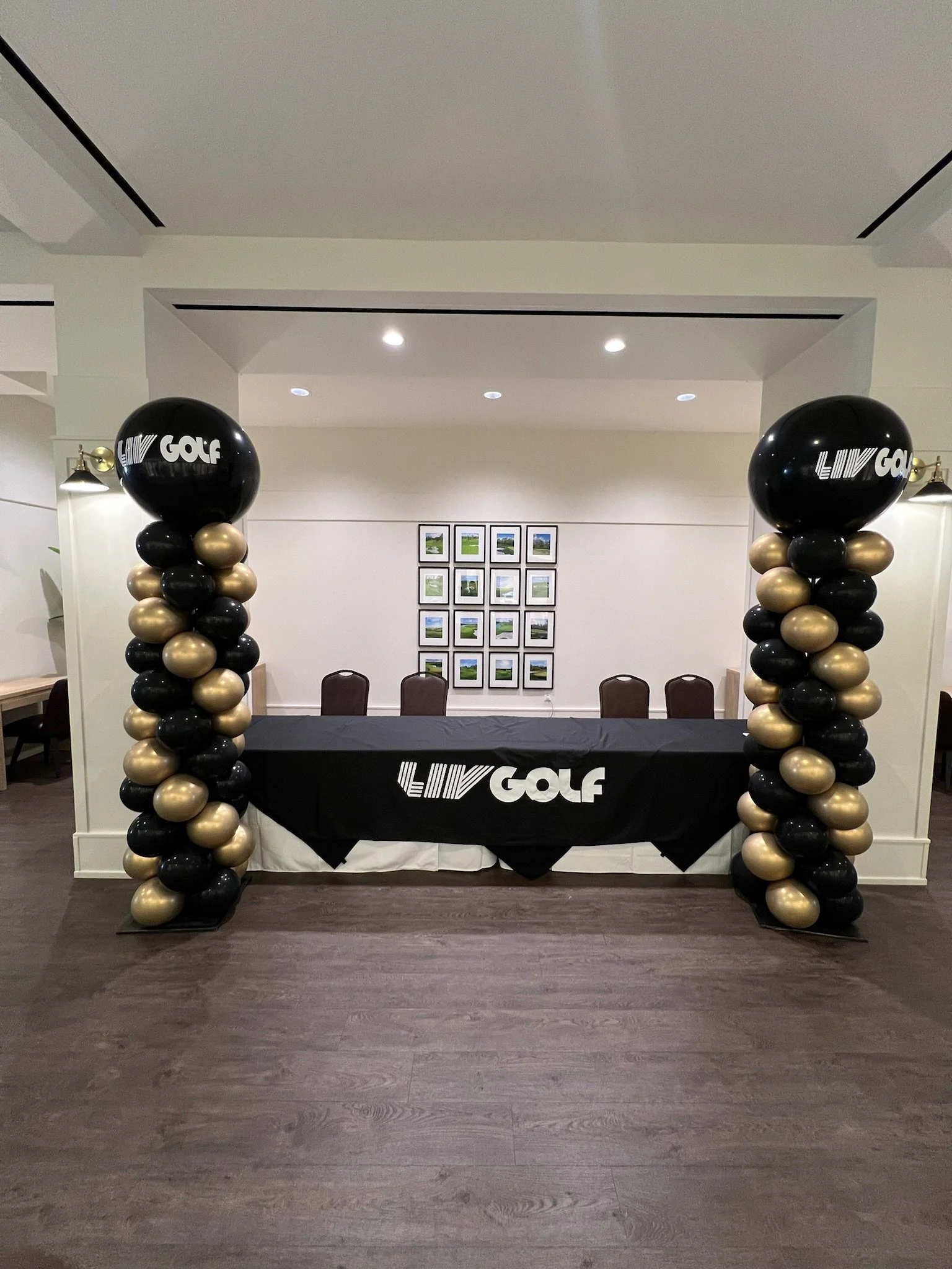Balloon arch with black and gold balloons on either side of a table with LIV Golf logo, set in a decorated indoor space.