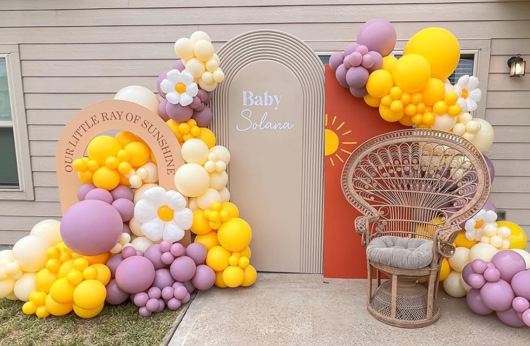 Decorative backdrop with balloons in yellow, purple, and white, featuring a sign that reads "Baby Solana" and a peacock-shaped chair.