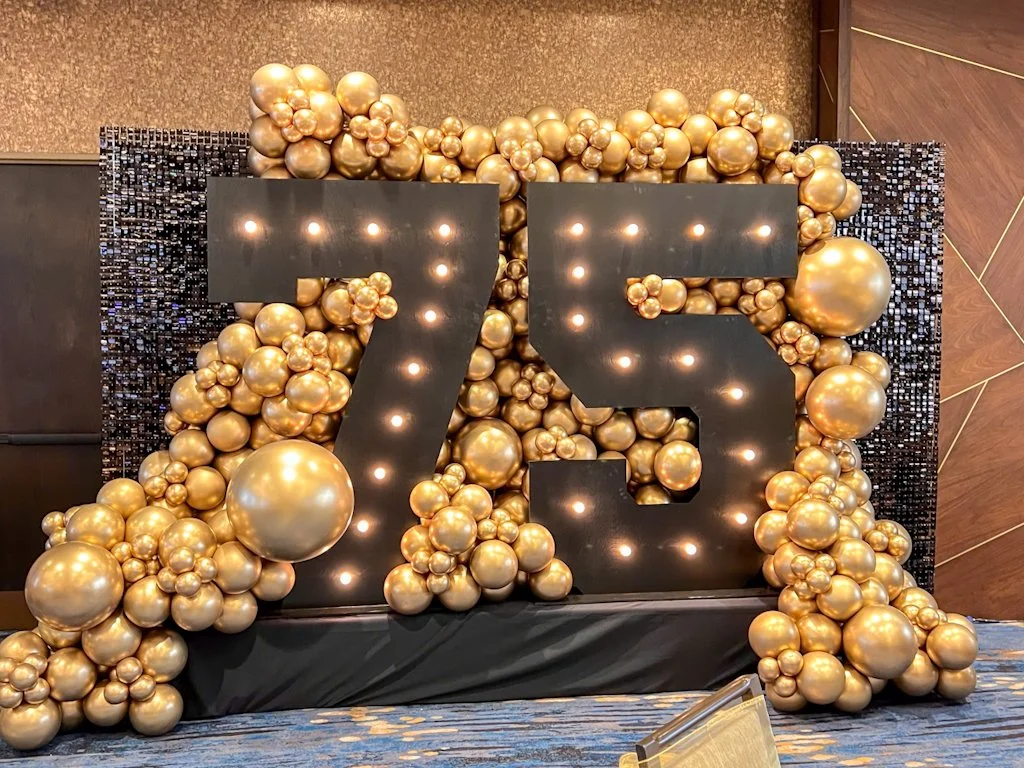 Large illuminated number 75 surrounded by gold balloons at a celebration or event.