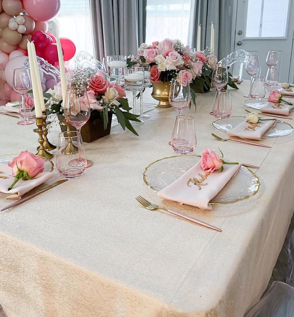 A decorated dining table with pink and white floral arrangements, gold and pink glassware, gold utensils, and pink rose napkins. There are tall white candles and floating candles in glass holders. The table is set for a celebration or special event, 