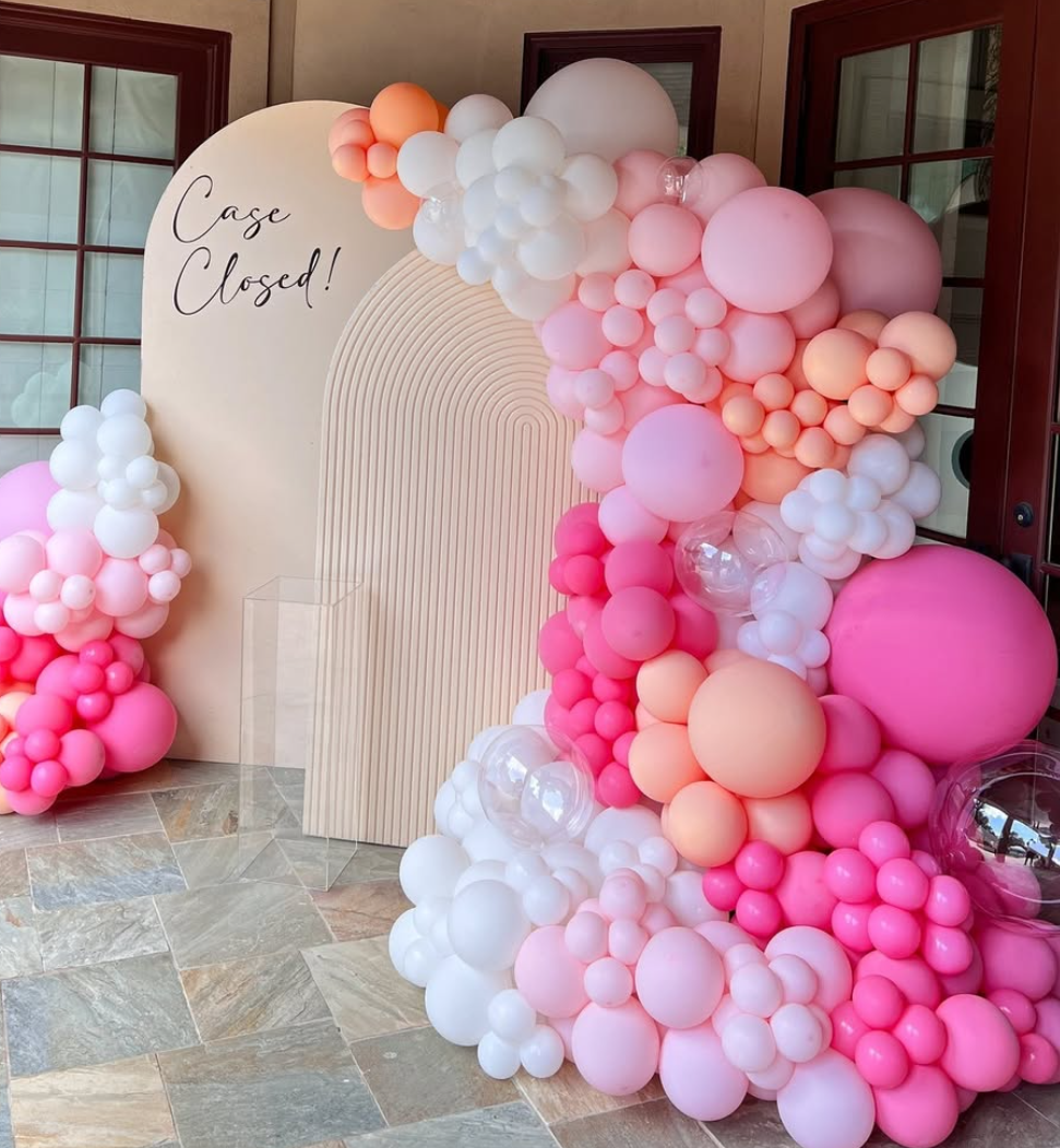 Decorative balloon arrangement in shades of pink, peach, and white surrounds a beige backdrop with the words 'Case Closed!' written on it, set on a stone tile floor near house windows.
