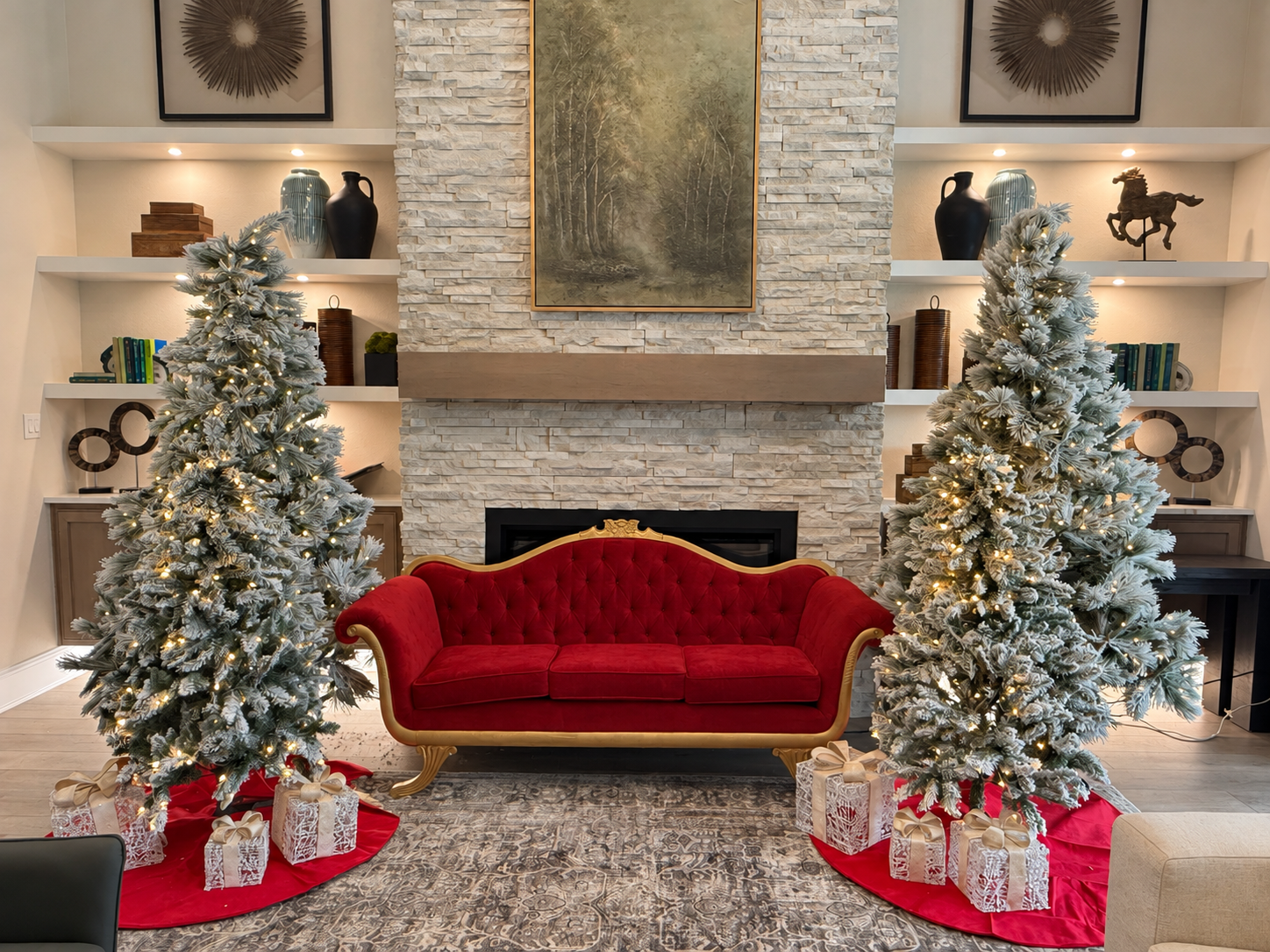 Red velvet sofa positioned between two illuminated, snow-covered Christmas trees in front of a stone fireplace.