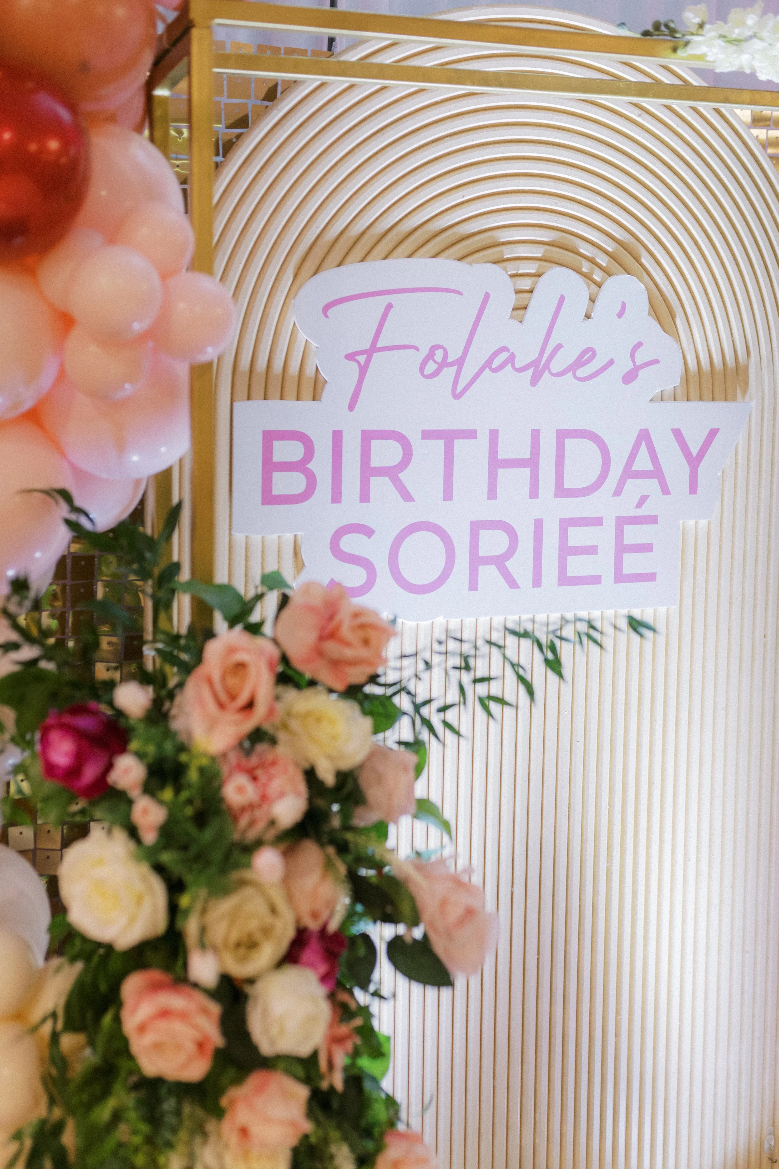 Decorative sign for a birthday celebration reading 'Folake's Birthday Soree' with a backdrop of textured gold and white fan decor, pink and white balloons, and a floral arrangement of pink, white, and cream roses.