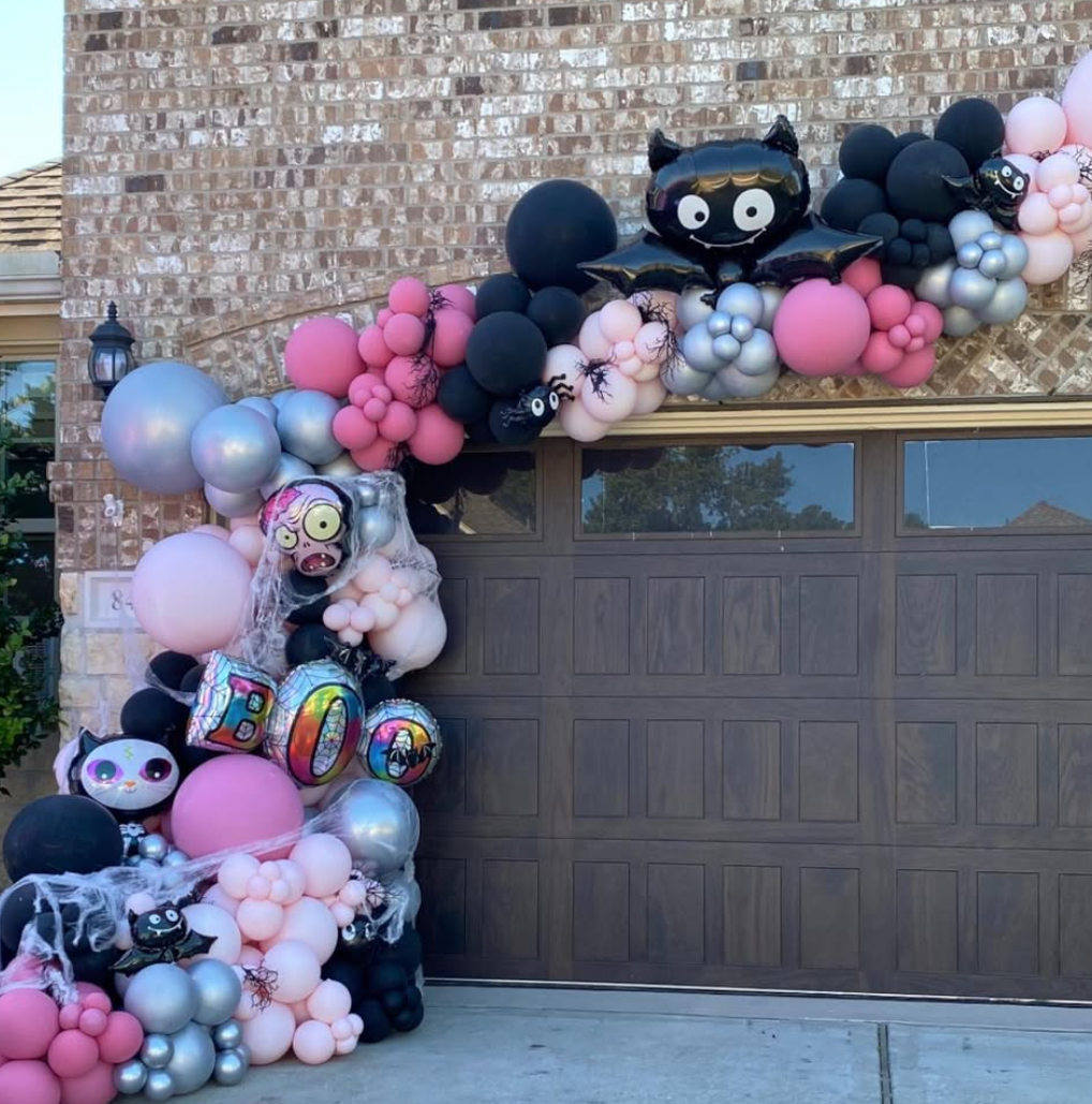 Halloween-themed balloon decoration with pink, black, and silver balloons, featuring cartoon zombie and bat balloons, cobweb decorations, and a large bat-shaped balloon, arranged around a garage door and on the side wall of a house.