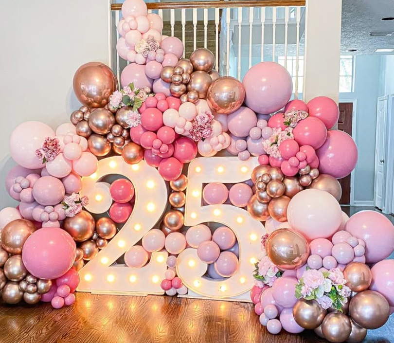 Decorative arrangement of pink, purple, gold balloons and flowers surrounding illuminated "25" for a celebration or birthday event.