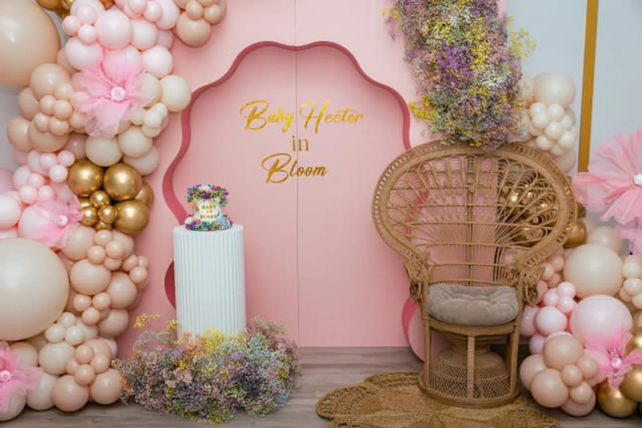A decorative backdrop for a baby shower, featuring pink and beige balloons arranged around a pink wall with the wording 'Baby Heair in Bloom' in gold. A white pedestal with a colorful cake is on the left, and a wicker peacock chair with pink and purp