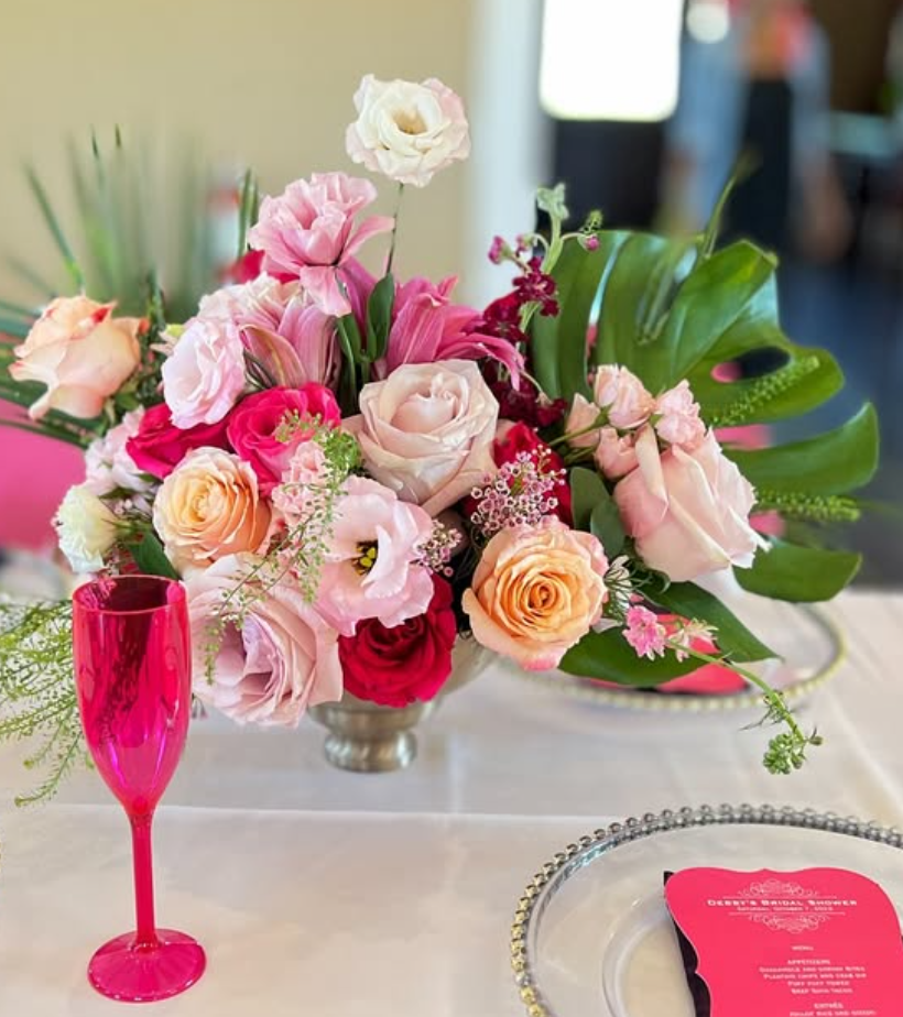 A floral centerpiece with pink, peach, and white roses, along with greenery, on a table with a pink champagne flute and a menu card.
