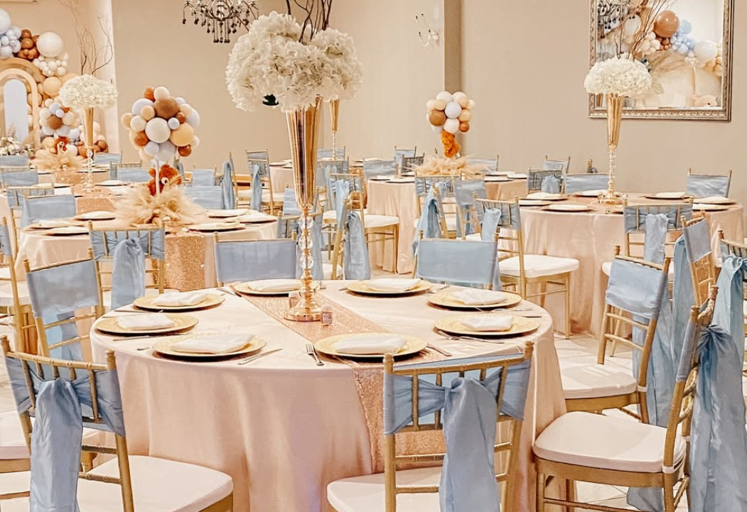 Elegant banquet hall decorated with tables covered in peach tablecloths, gold and blush accents, tall floral centerpieces with white flowers, and blue chair sashes, set for a celebration or wedding reception.