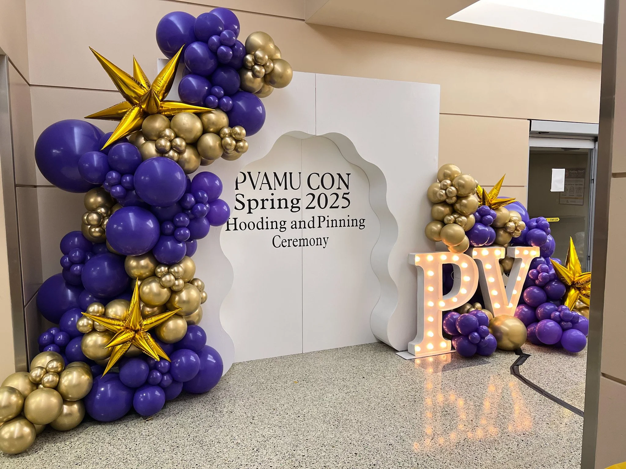Balloon display with purple and gold balloons, golden star-shaped balloons, and a lit-up sign with the letters 'PV' at a university spring 2025 hooding and pinning ceremony.