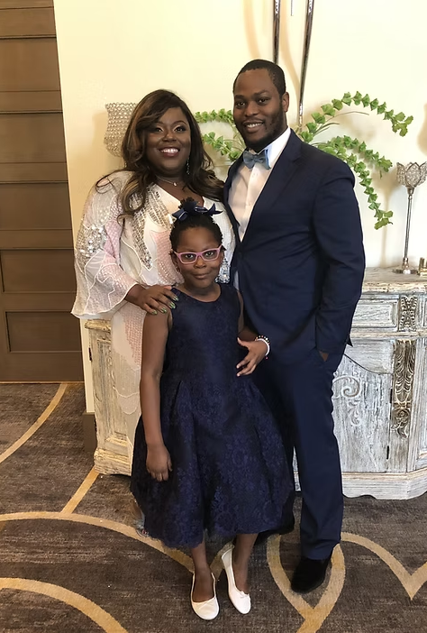A family of three posing indoors, with a woman, a man in a suit, and a young girl in a navy dress and pink glasses.
