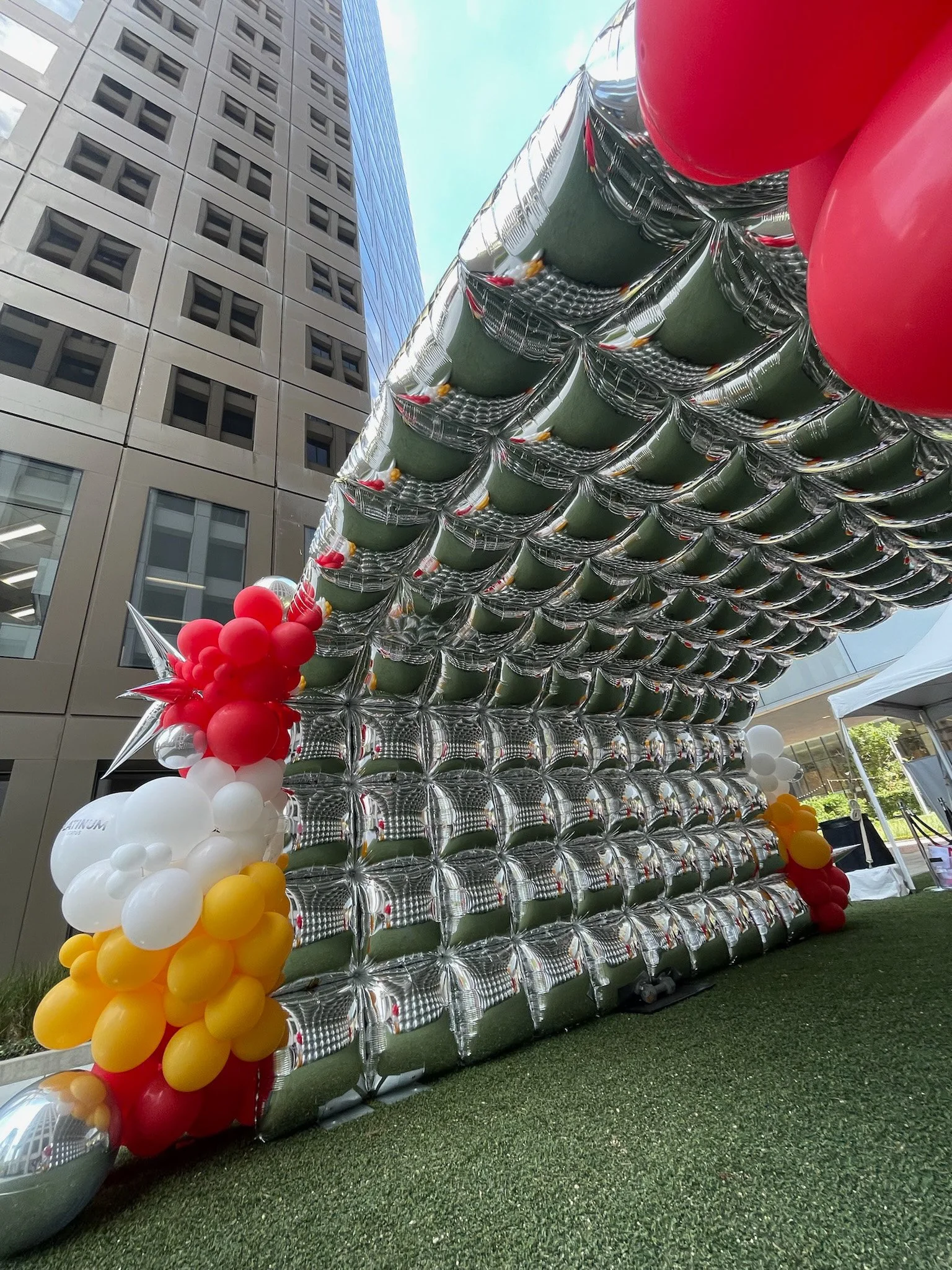 Large installation of metallic and colored balloons creating a sculpture or decorative structure outdoors, with modern buildings in the background.