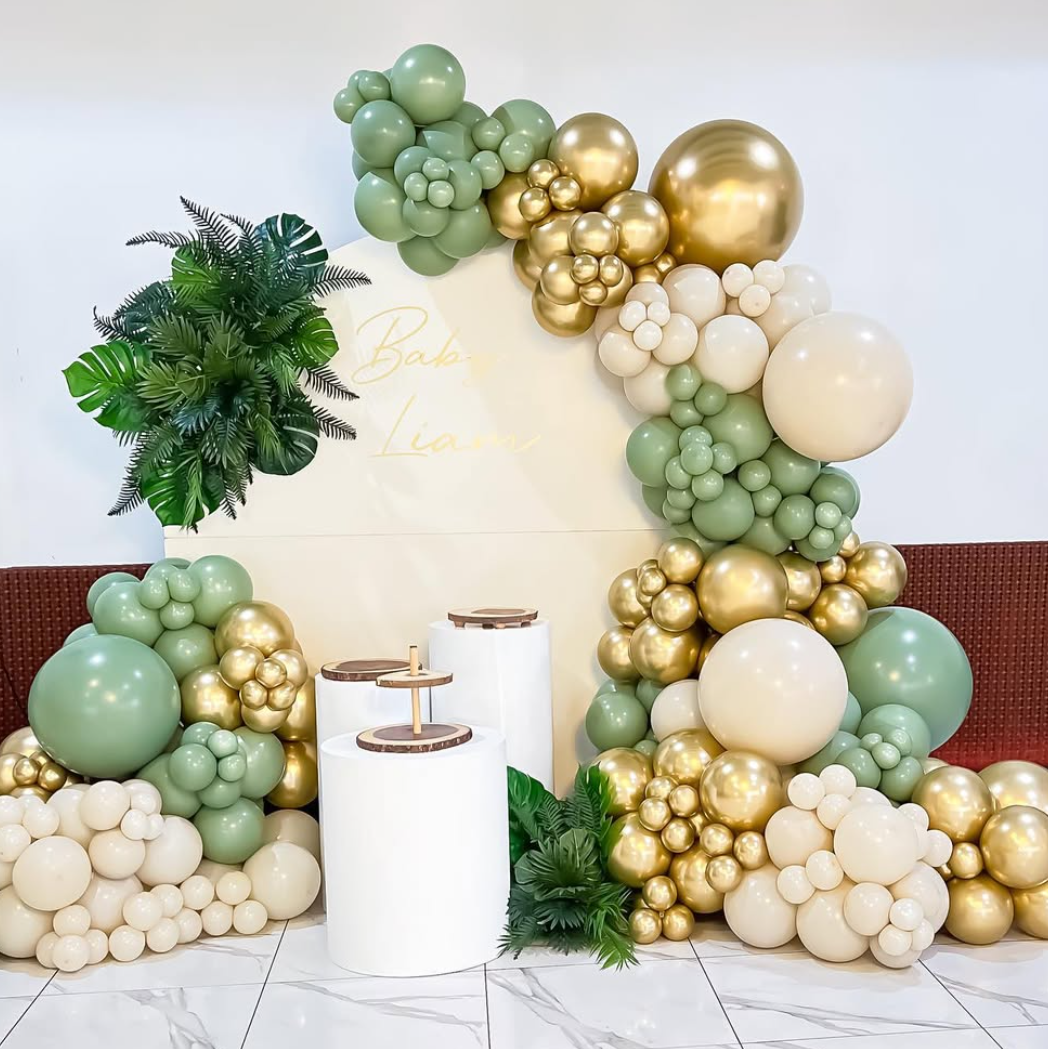 Decorative balloon arrangement with green, gold, and white balloons, a white backdrop with gold writing, green foliage, and white pedestal tables in front.