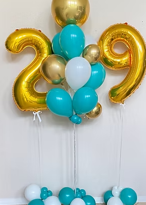 A birthday celebration display featuring large gold foil balloons shaped as the numbers 2 and 9, surrounded by blue, gold, and white balloons arranged in a cluster against a wall.