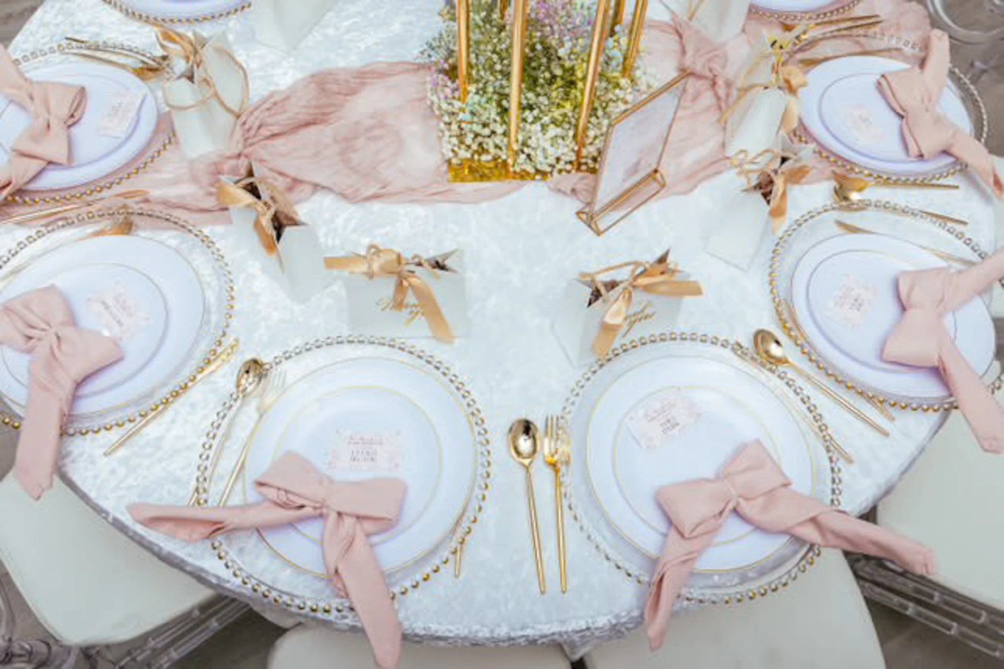 An elegantly set round table with six place settings, each with a white plate, pink napkin tied in a bow, gold utensils, and a decorative plate border. Centerpiece features a gold geometric frame filled with white flowers and baby's breath, with pink