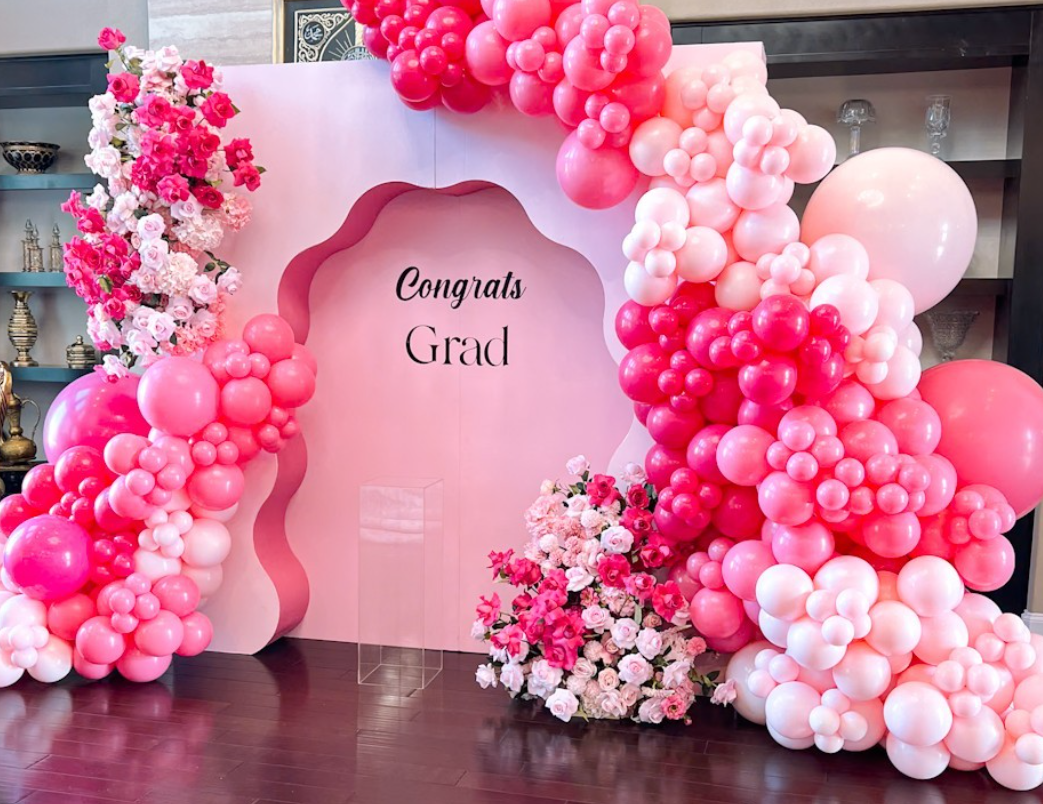 Pink and white balloon arch with floral arrangements and a pink backdrop that says "Congrats Grad" in black script.