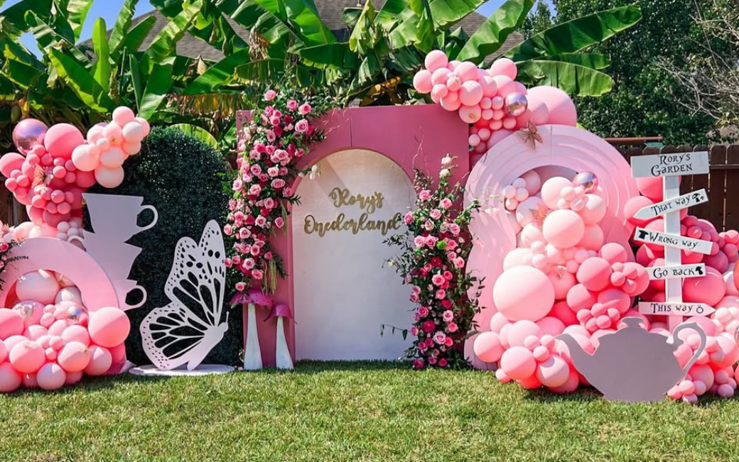 Decorative garden backdrop with pink balloons, flowers, butterfly and swan cutouts, surrounded by tropical banana trees, with a sign reading 'Rory's Wonderland' and a signpost with directions.