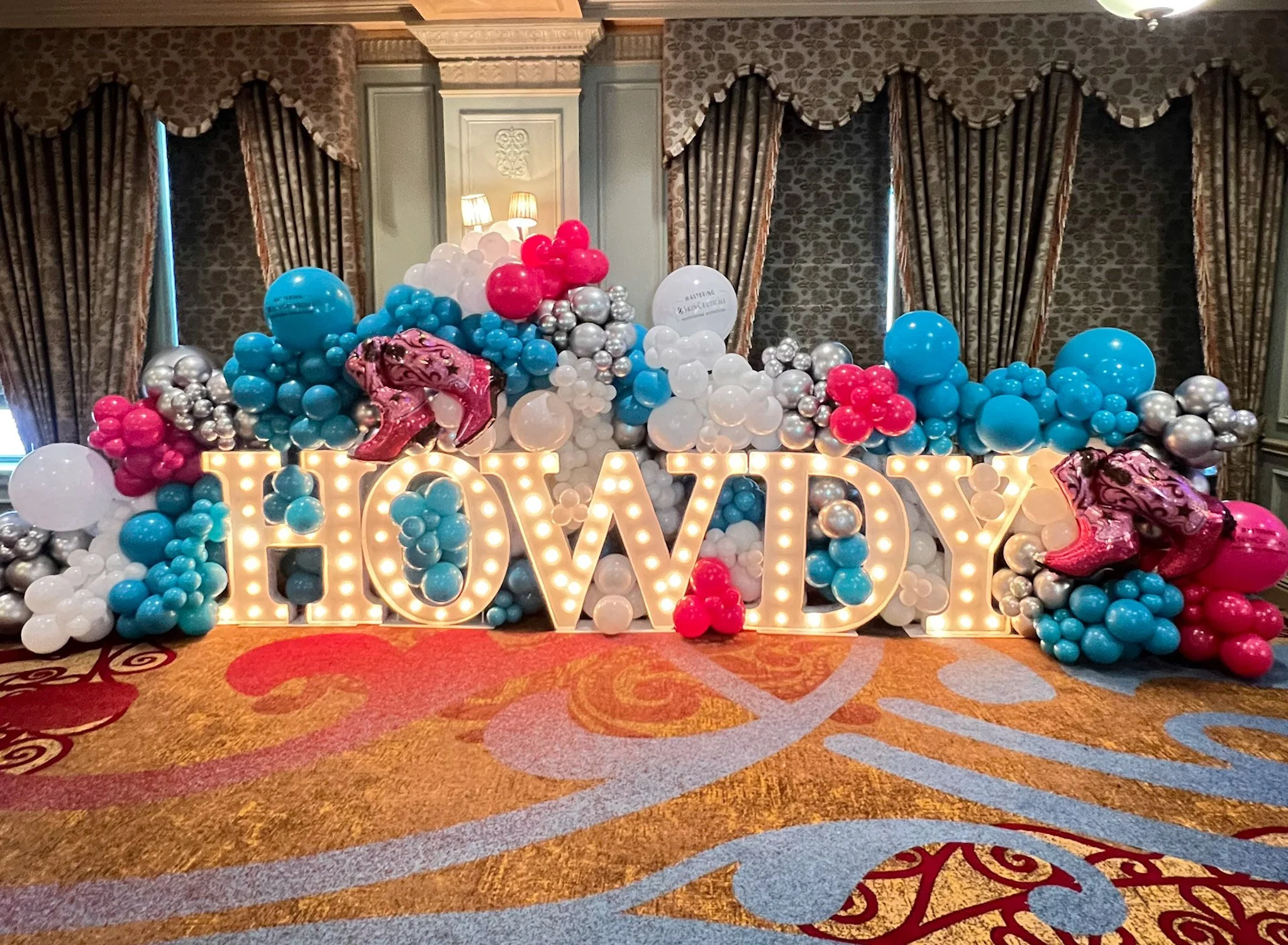 Decorative balloon display with pastel blue, pink, white, and silver balloons behind a large illuminated "HOWDY" sign, with pink cowboy boots on each end, set in a room with patterned curtains and a carpeted floor.