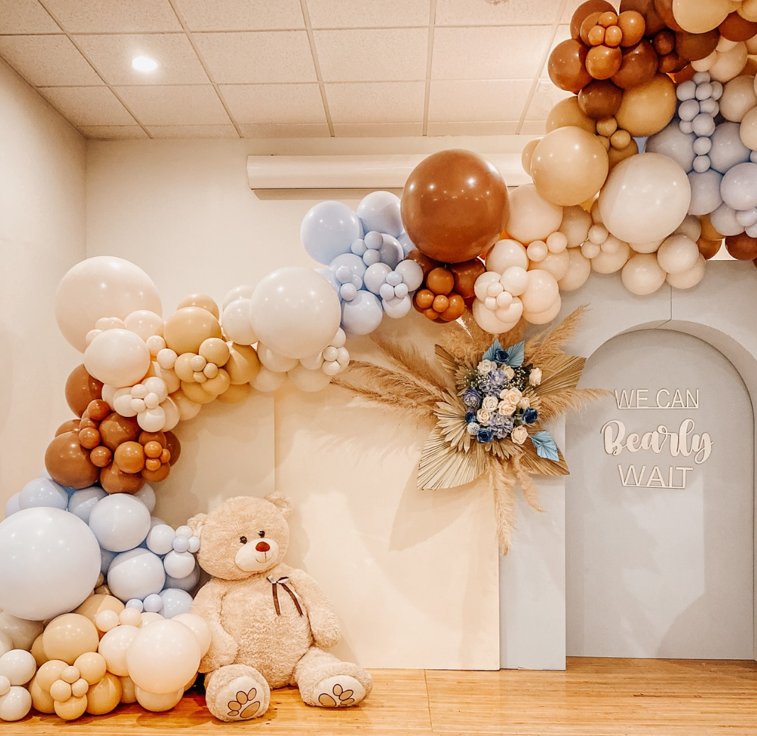 Decorative balloon arrangement in pastel colors with a large teddy bear, dried flowers, and a sign that says 'We can bear-ly wait' in a room with wooden flooring.