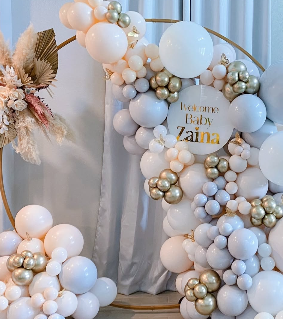 Decorative balloon arrangement with white, gold, and pastel balloons, gold butterflies, and a sign that reads "Welcome Baby Zaina" at a celebration.