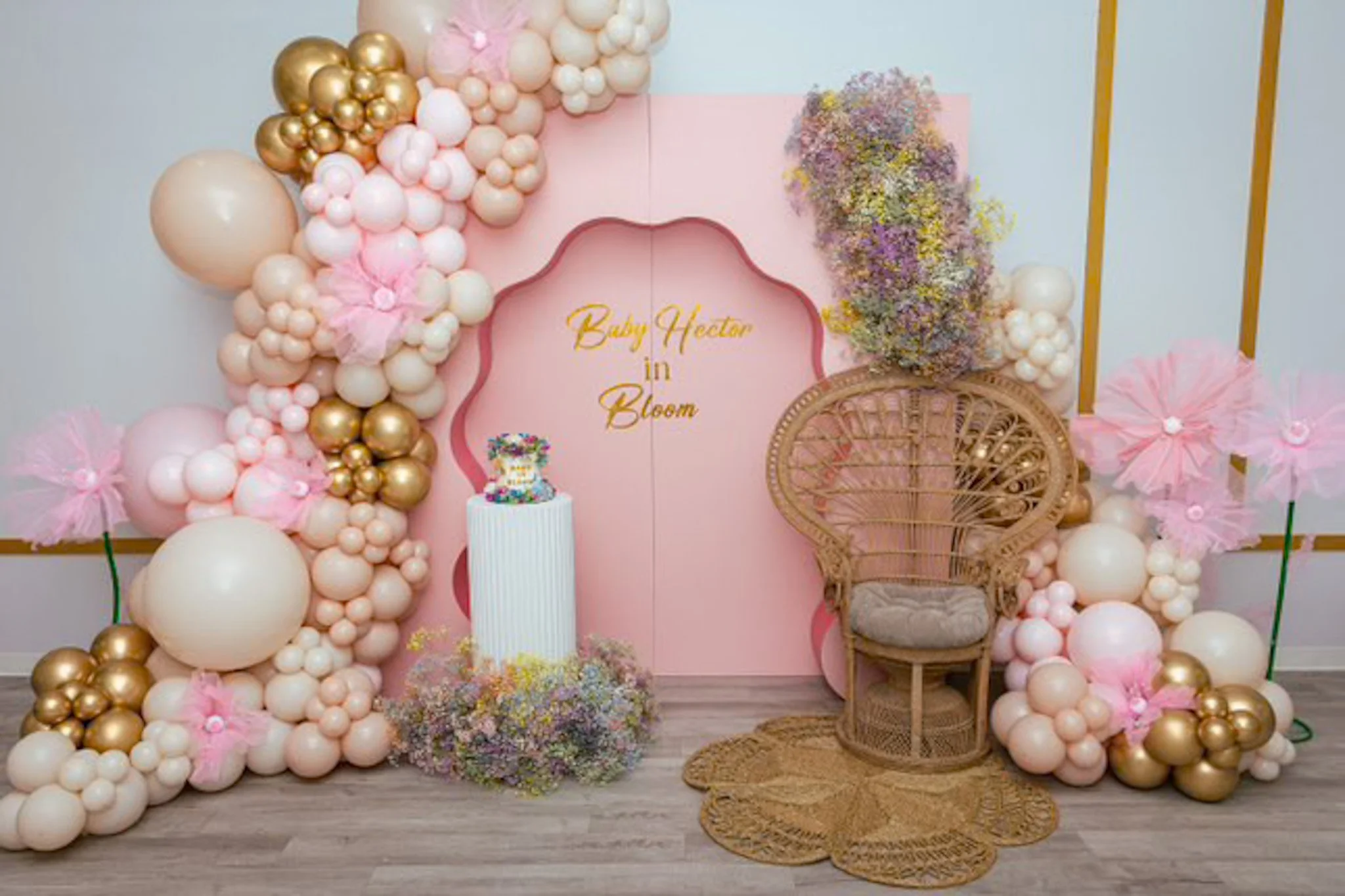 Decorative setup with pink and gold balloons, a pink backdrop with the text 'Baby Hector in Bloom,' a wicker chair, a small cake, and pink floral decorations, indicating a celebration or baby shower.
